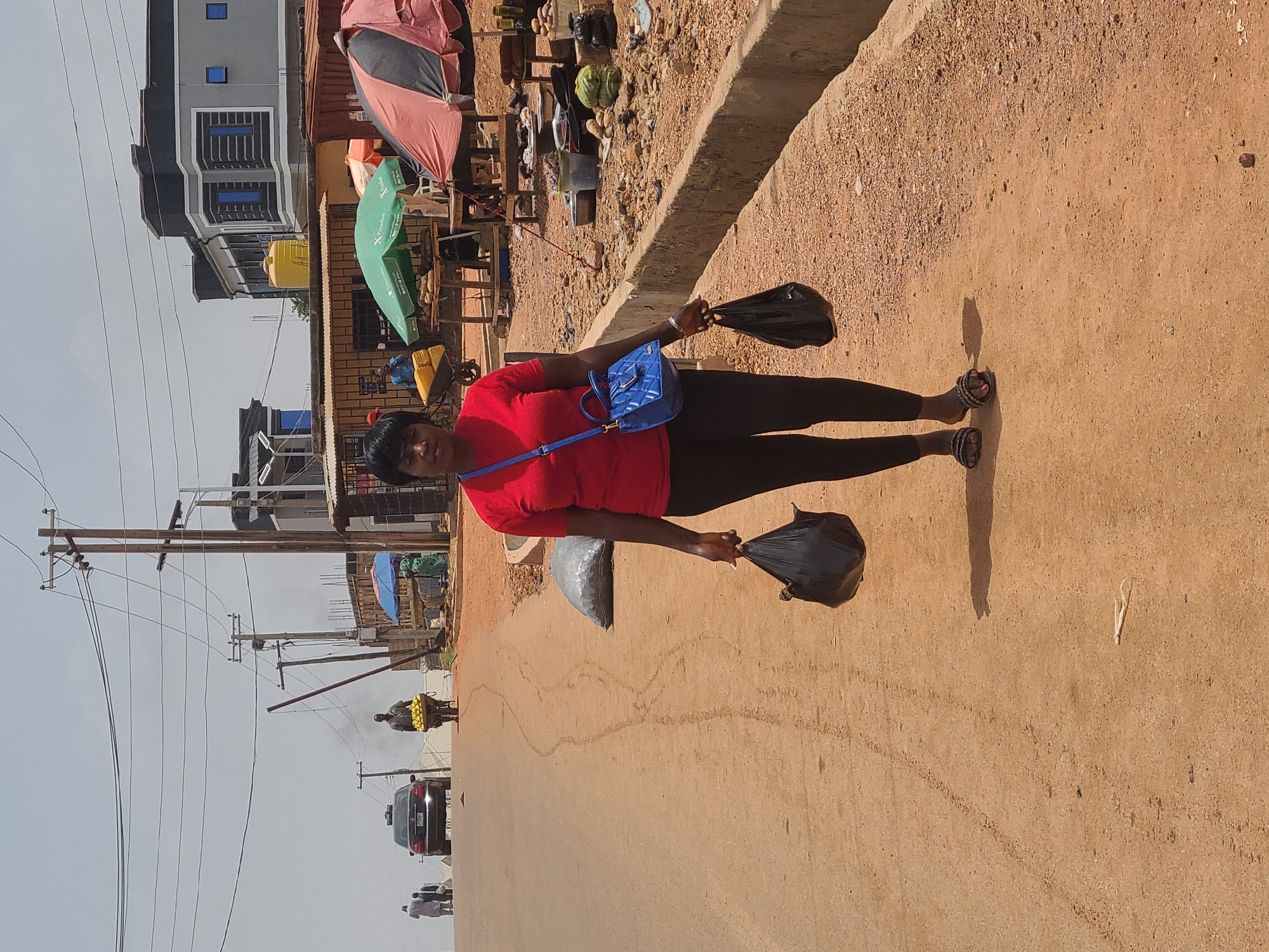 A woman stands on a street in Jos, Nigeria, holding black plastic bags in each hand. She is wearing a red shirt, black leggings, and sandals, with a blue purse slung across her body. Behind her, there are roadside market stalls with umbrellas, a building with multiple windows, utility poles with wires, a car, and people. The ground is unpaved.