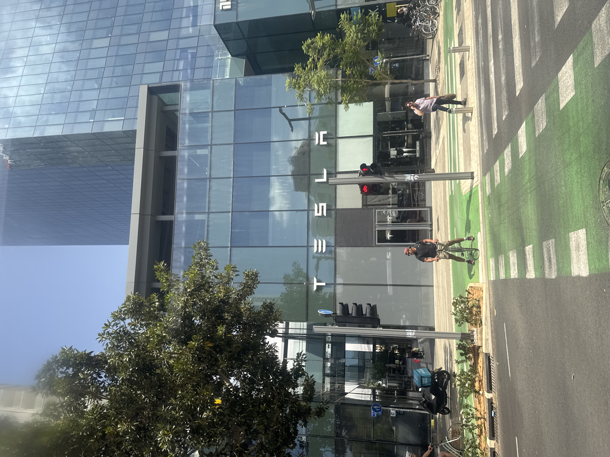 The image captures a street scene in Tel-Aviv, Israel, featuring a Tesla store. The store's facade is mostly glass. The street has a pedestrian crossing painted with green stripes. A man on a bicycle is on the zebra crossing. A woman is walking in the same direction near the store. Traffic lights are visible. A tree is in the foreground. A large, modern building is visible behind the Tesla store. Parked scooters are nearby.