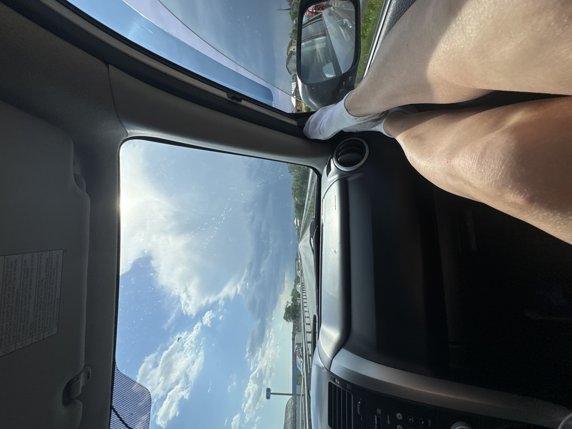 A person's legs are propped on the dashboard of a car while it is in motion on a road in Kitsjanovo, Russia. The sunny sky and the car's dashboard and interior are visible.