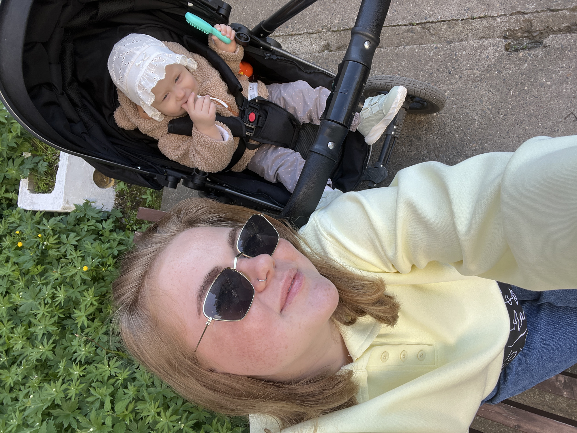 The image shows a woman and a baby in a stroller, potentially taken in Chaikovsky, Russia. The woman, wearing sunglasses and a yellow shirt, is lying on a surface covered in greenery. The baby is secured in the stroller, wearing a bonnet and a brown jacket. The stroller is black with grey detailing and appears to be on a paved surface. The overall setting suggests an outdoor location, possibly a park or residential area.