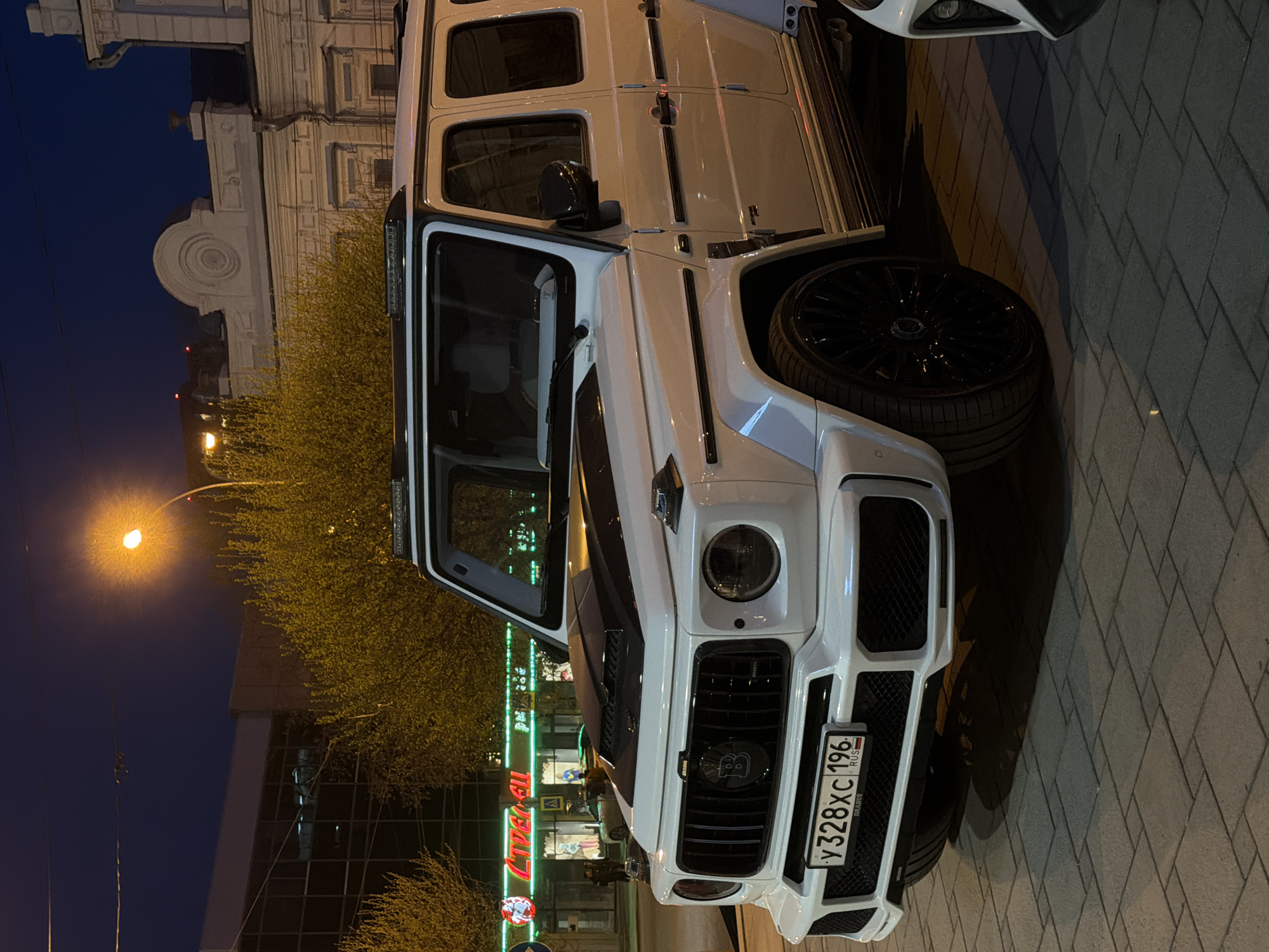 The image shows a white Mercedes-Benz G-Class Brabus parked on a paved sidewalk in front of a brightly lit commercial building in Yekaterinburg, Russia. The car has a distinctive front grille with a prominent "B" logo and a Russian license plate that reads "У 328 ХС 196 RUS". The building across the street has several store signs, some of which are illuminated with red and green lights. There are trees to the side of the building with yellow flowers or leaves. A streetlamp is visible in the upper left portion of the image. The sky is dark, suggesting it is either late evening or night.