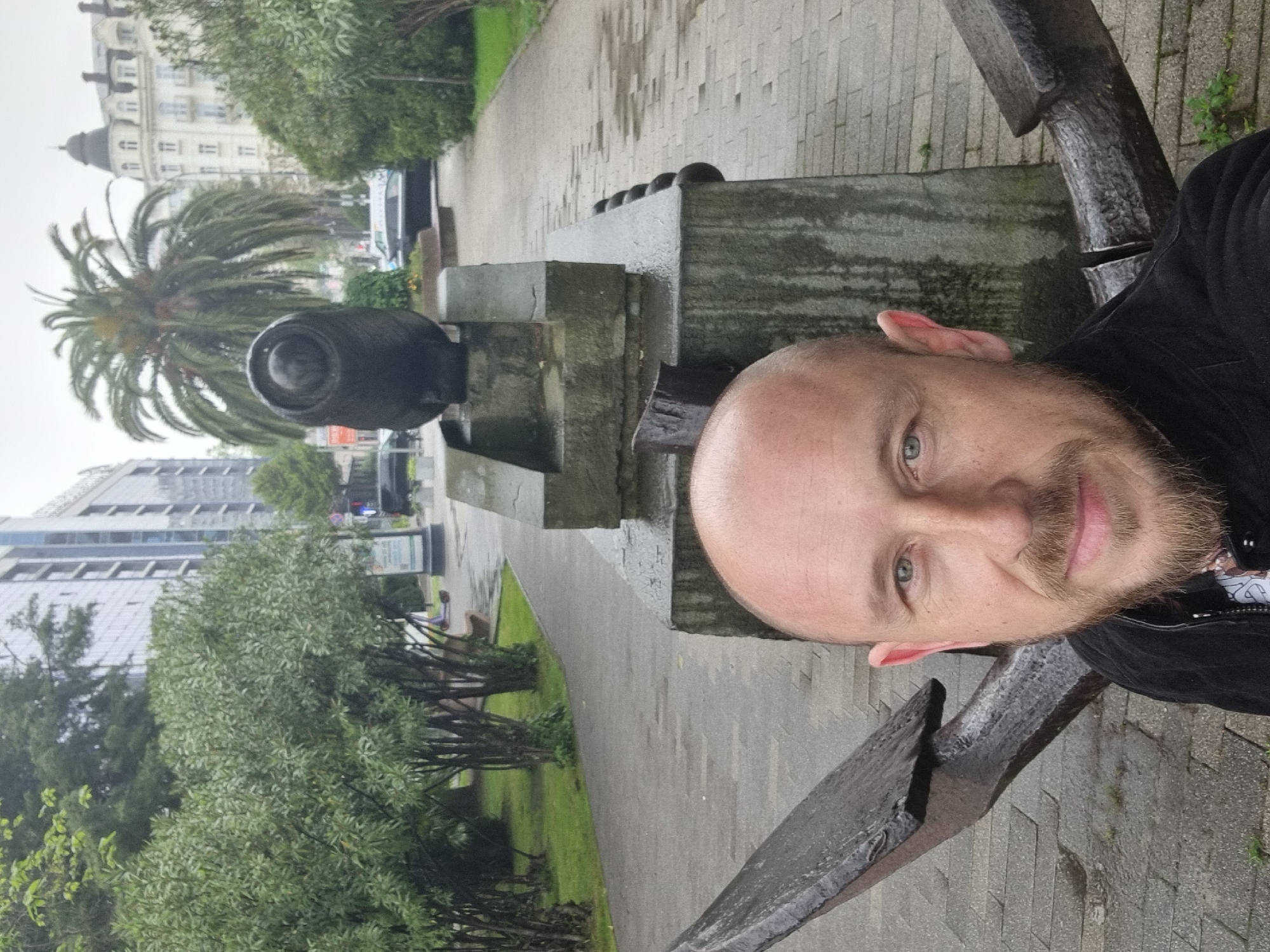 A medium shot shows a man taking a selfie. He is a white male with a bald head and short beard. He is positioned to the right of the frame, leaning against a large black metal anchor. The anchor is part of a monument or memorial. The background consists of a paved walkway, green lawn, and various trees. In the distance, there are buildings and palm trees. This scene is likely in the city of Sochi, Russia based on the type of monument and urban landscape. The lighting appears overcast.
