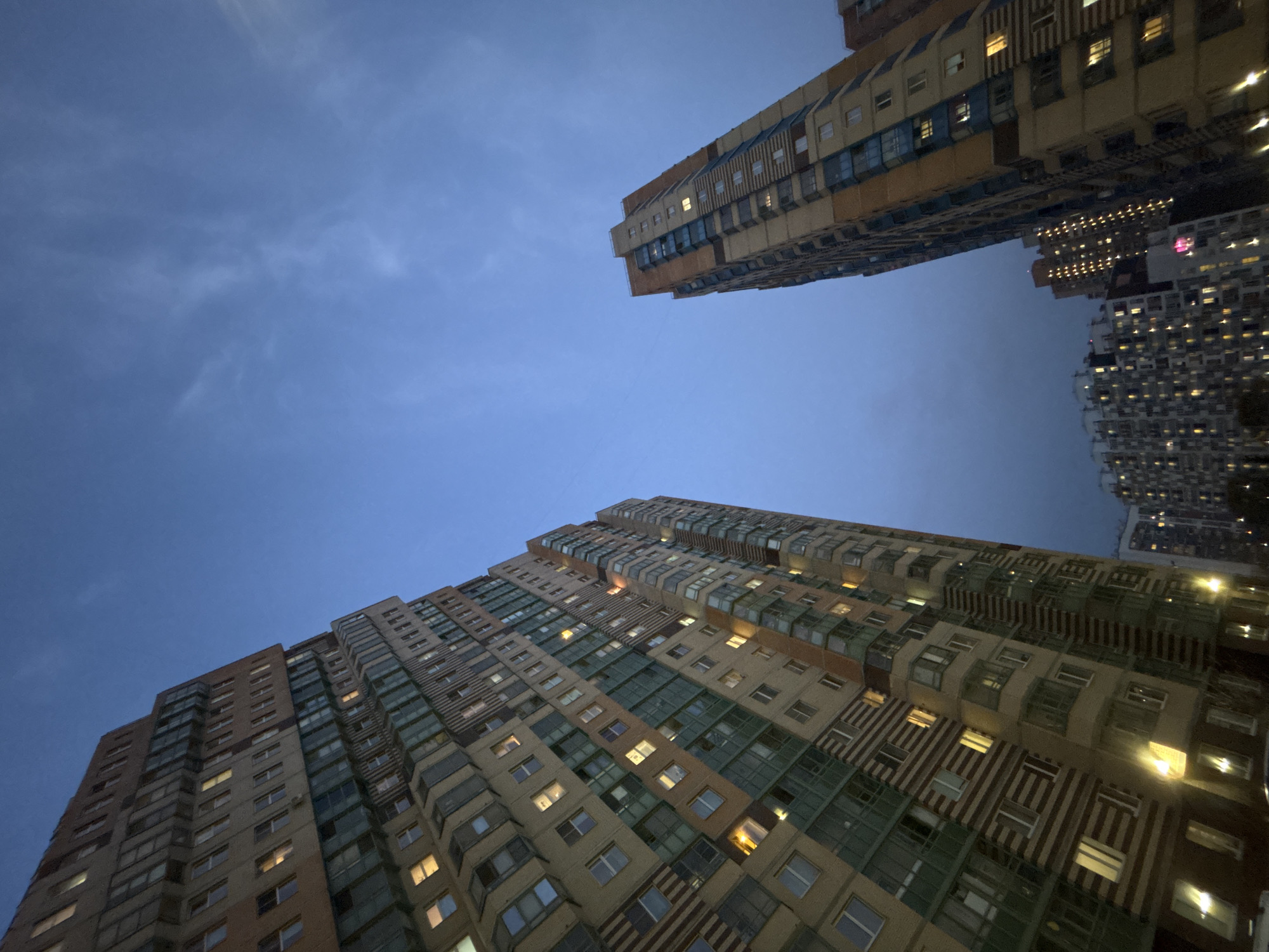 The image depicts a low-angle view of high-rise residential buildings in Kudrovo, Russia, under a cloudy evening sky. The buildings are densely populated, with many illuminated windows suggesting residential activity. The architectural styles appear modern, featuring a mix of colors and designs on the facades. The overall environment conveys an urban residential setting at dusk or nightfall.