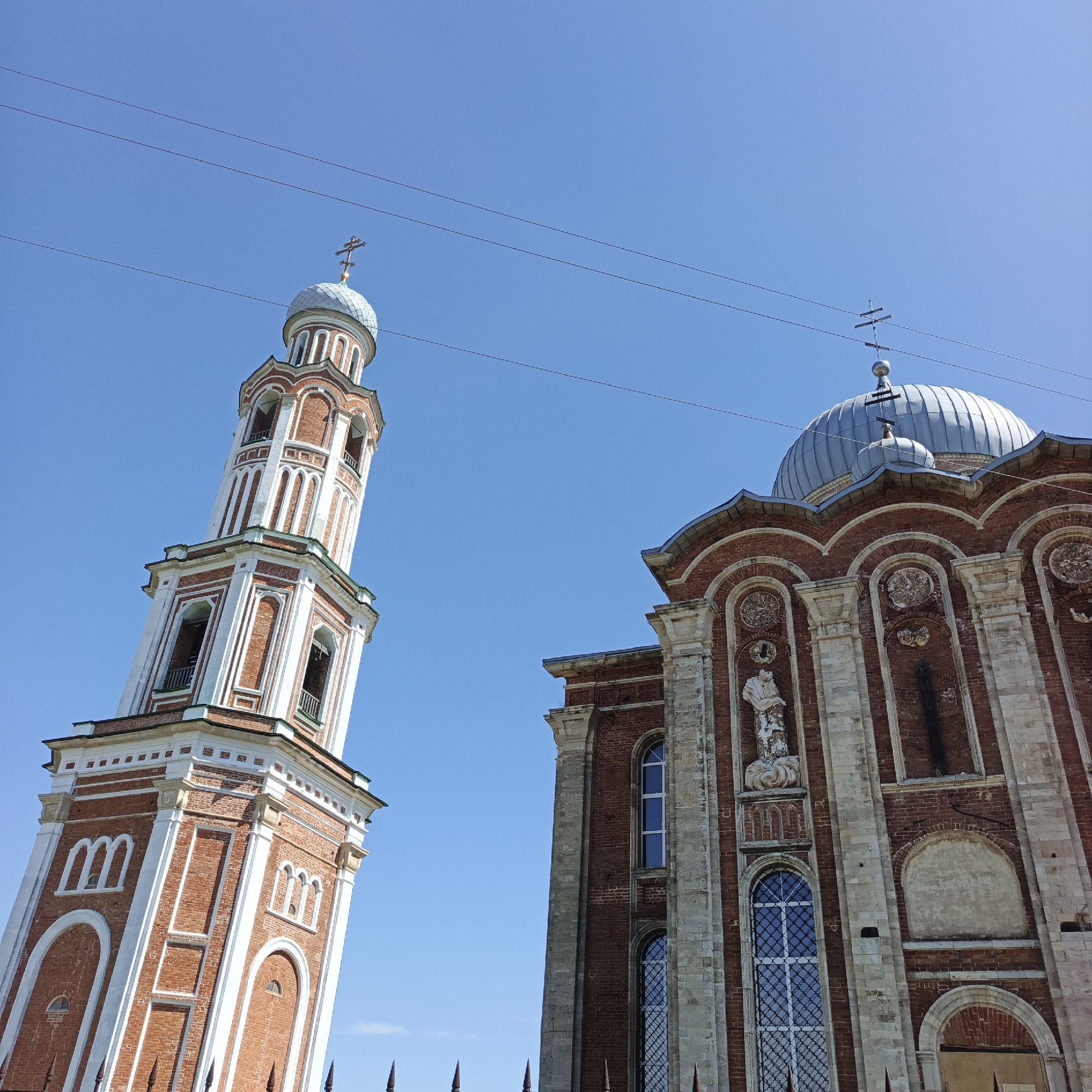 The image shows a low-angle view of the bell tower and main building of the Church of the Resurrection of Christ in the village of Tugustemirovo, Kirzhalsky District, Sverdlovsk Oblast, Russia. The bell tower is a tall, slender structure with red brick and white trim, featuring multiple levels with arched windows and decorative elements. It is topped with a light blue dome and a cross. To the right of the bell tower is the main building of the church, also constructed of red brick with lighter stone accents. This building features a large, gray, ribbed dome topped with a cross, and its facade is adorned with arched windows, niches, and decorative carvings. The sky above is clear and bright blue, with power lines crossing horizontally across the frame. The image captures the architectural grandeur of the religious complex under a sunny sky.