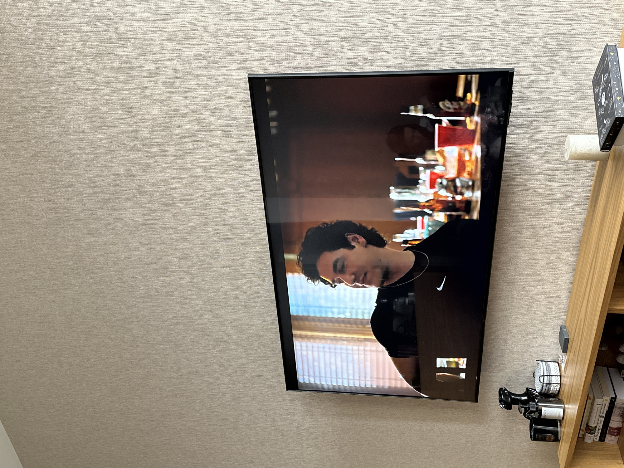 The image shows a wall-mounted television displaying a scene with a man wearing a black shirt with a white Nike logo. The background on the screen suggests a bar setting with bottles and glasses. To the right of the TV is a wooden shelf unit. The wall behind the television is a light beige color with a textured surface. Without additional information, the location in Yerevan, Armenia, cannot be confirmed.