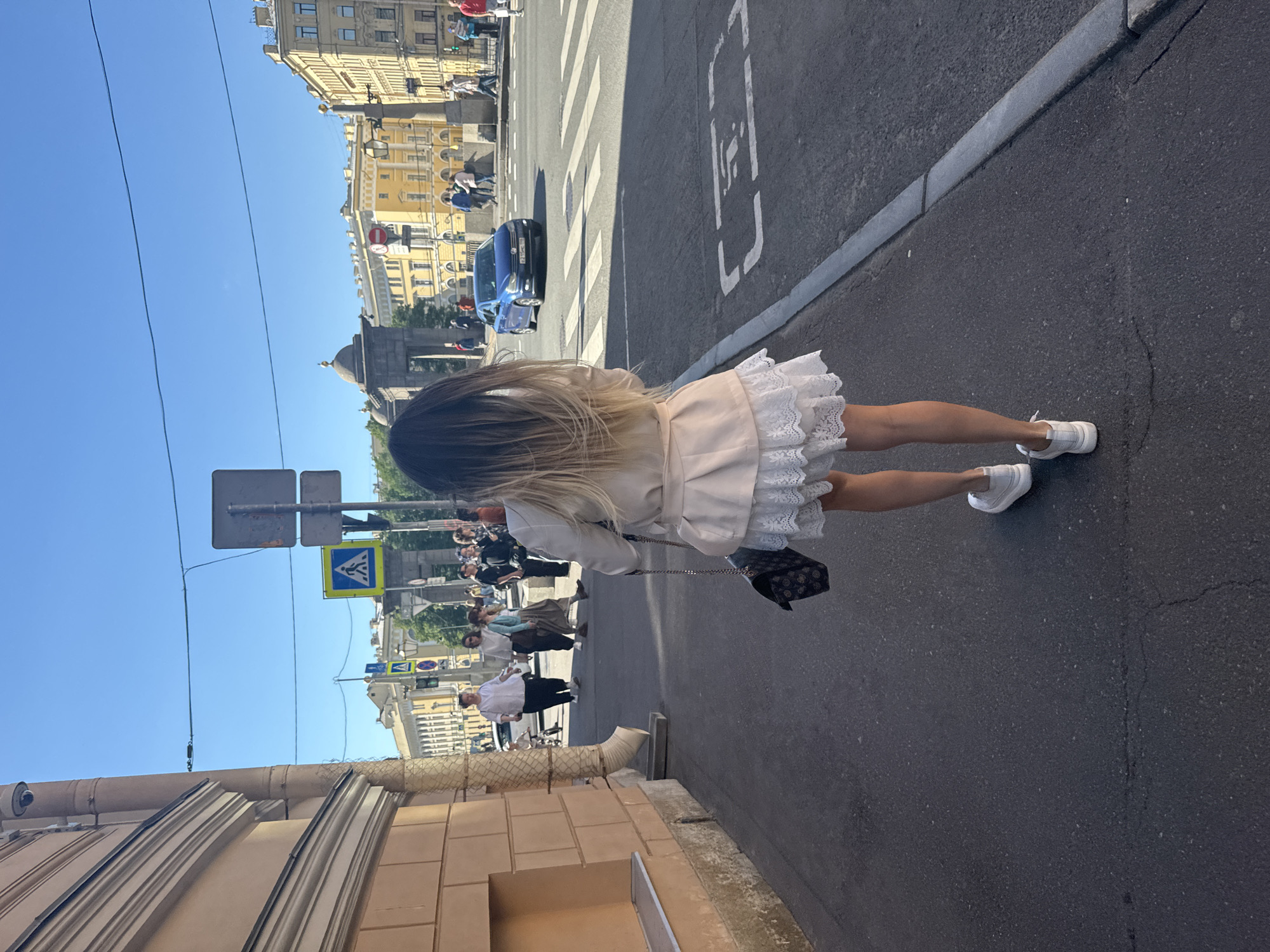 The image shows a woman walking away from the camera on a sidewalk in Saint Petersburg, Russia. She is wearing a light-colored jacket over a white lace dress and white sneakers. She is carrying a dark patterned handbag. The sidewalk is asphalt, and there is a curb separating it from the street. Across the street, there are buildings, a crosswalk, and traffic. The sky is blue and clear. The architecture of the buildings suggests a historical European city, consistent with Saint Petersburg. There is a pedestrian crossing sign visible in the distance, and a blue car is crossing the street. The overall scene depicts a sunny day in a bustling city.