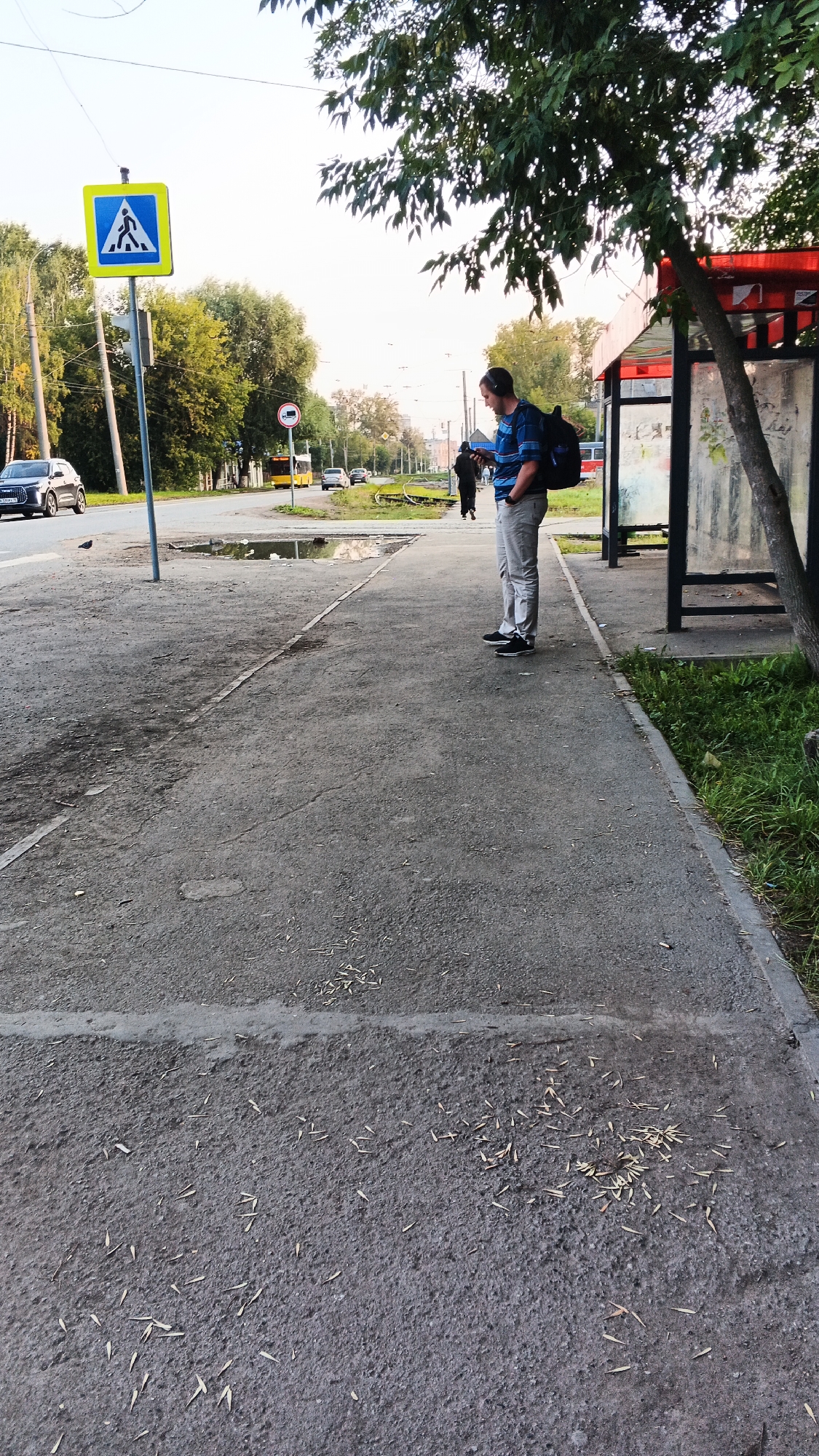 This is an eye-level, full shot of a man standing at a bus stop in Izhevsk, Russia. The man is wearing a blue and black striped t-shirt, grey pants, and black sneakers. He has headphones on and is looking at his phone. He is also carrying a black backpack. A second, darker figure, likely a woman based on clothing, is walking away from the bus stop in the background. The bus stop has a red roof and glass panels. A pedestrian crossing sign is visible in the background. The man is standing on a sidewalk next to a road with a few cars and a yellow bus. There are trees and other buildings visible in the background. The ground is paved with asphalt and there are some puddles on the sidewalk. The sky is clear and overcast.