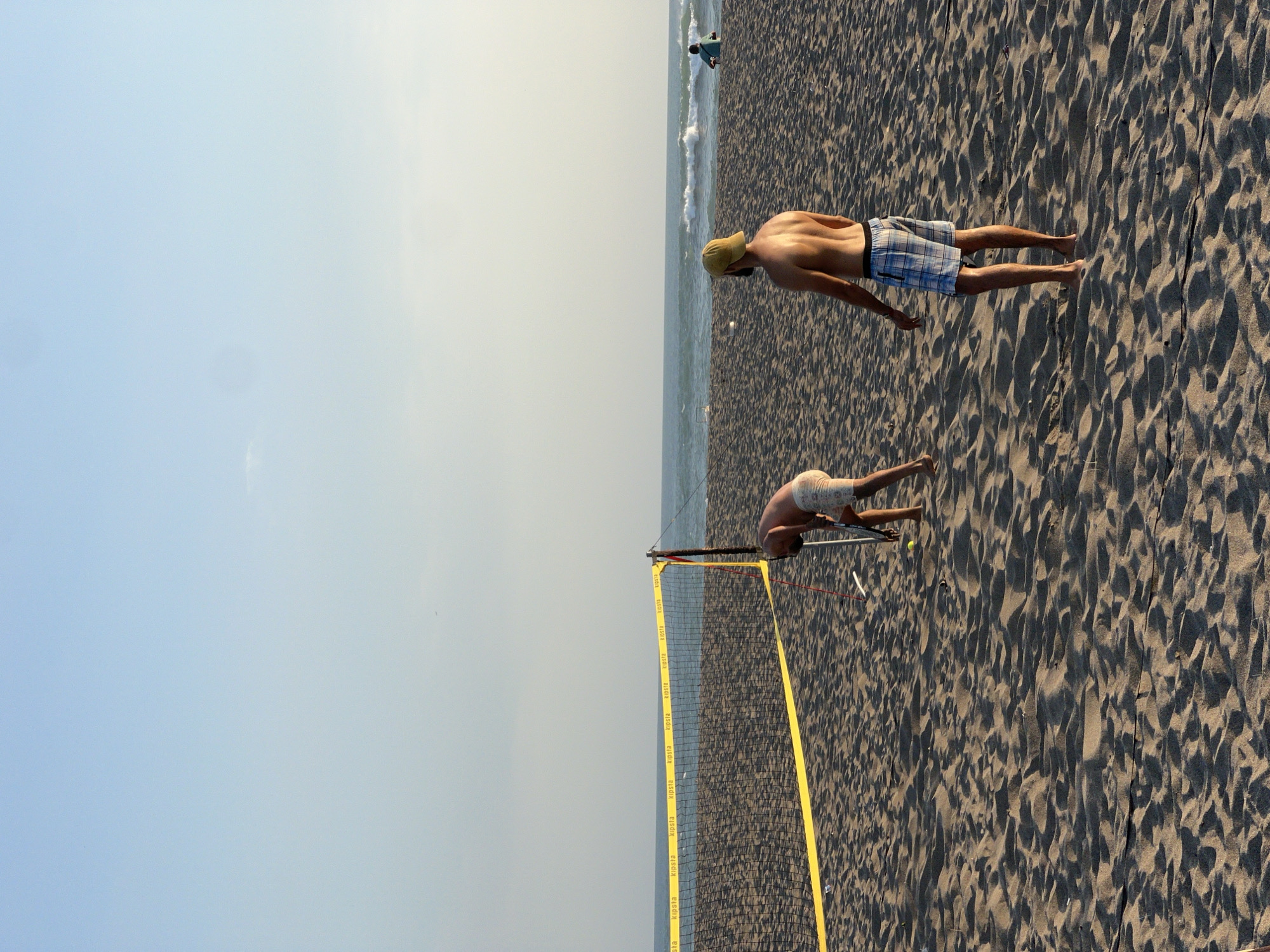 An outdoor, medium-shot captures two men playing on a sandy beach in North Kuta, Indonesia. The sky is clear and pale blue, with a faint hazy horizon where the water meets the sky. A volleyball net, with a yellow boundary tape, is set up in the sand, dividing the playing area. One man, wearing a yellow cap and checkered shorts, is standing with his back to the camera, facing the net and the ocean. He appears to be waiting or observing. To the left of the net, another man, wearing patterned shorts, is bent over, holding a racket and appearing to interact with a yellow ball on the sand. The sand is textured with ripples and shadows, indicating it is a beach. In the distance, the ocean has moderate waves, and a few people are visible in the background, one sitting and another standing further down the beach. The lighting suggests it is late afternoon.