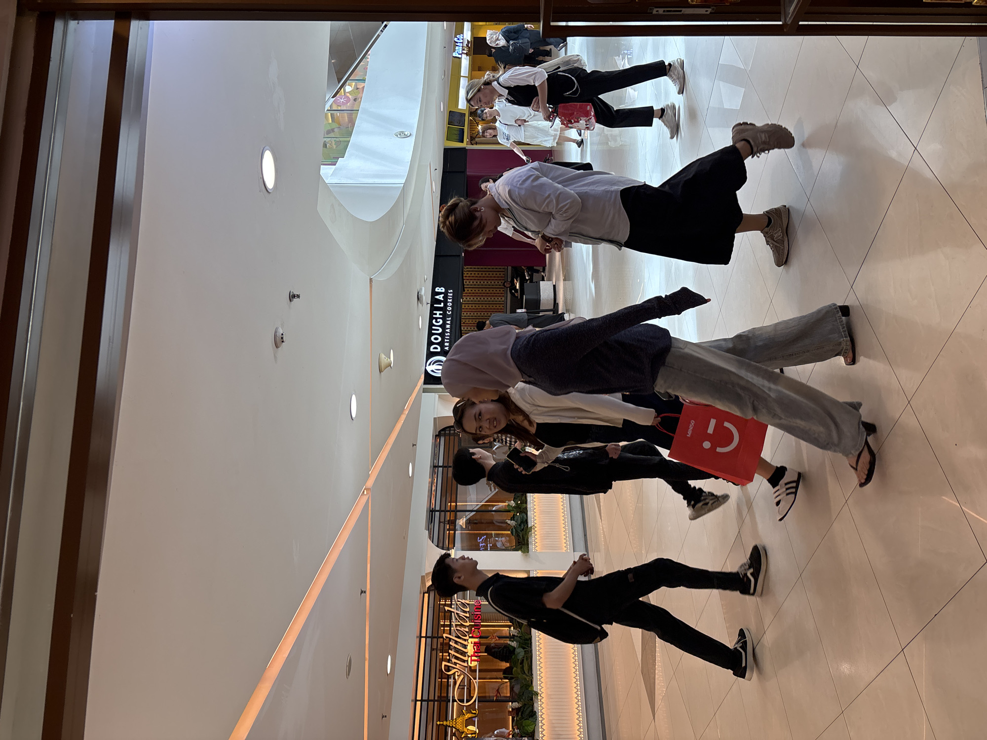 In a bright, modern indoor shopping mall located in West Jakarta, Indonesia, a group of people are seen walking on a polished white tiled floor. The mall features illuminated ceiling panels and decorative lighting. On the left, a young man in a black t-shirt and pants walks with a black sling bag over his shoulder. Next to him, a woman in a white long-sleeved top and black skirt is talking to another woman in a light purple hijab and a dark top. Further to the right, a woman in a white button-up shirt and a black skirt is looking at her watch, while another woman in a white t-shirt and a black vest with a white chain detail walks with a red shopping bag. In the background, store signs are visible, including "Jittlada Thai Cuisine" with ornate gold lettering and "DOUGH LAB ARTISANAL COOKIES" with a black background. The overall scene depicts casual activity within a commercial setting.