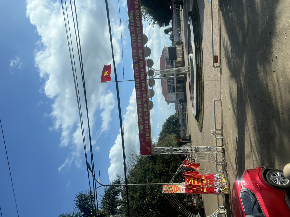 The image depicts a public square or town center in Trung Hồ, Vietnam, under a partially cloudy blue sky. In the foreground on the left, a red Nissan Kicks SUV is partially visible. The midground features a white metal archway entrance adorned with multiple small red Vietnamese flags. A prominent red banner with yellow Vietnamese text and illustrations of doves and gears is stretched horizontally across the archway, reading "STRONGLY AROUSE THE SPIRIT OF SELF-MASTERY, SELF-CONFIDENCE, SELF-RELIANCE, SELF-RESILIENCE, NATIONAL PRIDE, ASPIRATION TO DEVELOP THE COUNTRY IN THE NEW ERA." To the right, a large circular dry fountain or planter contains a modern, multi-branched sculpture with spherical elements. A "No Parking" sign in Vietnamese is visible on the paved ground near the fountain. In the background, a two-story building with a red-tiled roof and white walls stands at the far end of the square. To the right of this building, a tall flagpole displays a Vietnamese national flag (red field with a yellow star). The square appears empty of people. Palm trees and other foliage are visible on the left and in the distant background. Multiple dark overhead wires cross the upper portion of the frame.
