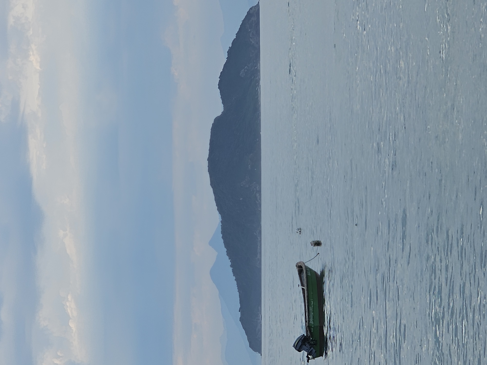 A long-distance shot shows a green boat floating on the water. In the background, there is a mountain with its shadow reflecting onto the water. The sky is light blue with some clouds. This is likely a view of the ocean near the city of Armação dos Búzios, Brazil, a beach resort city.