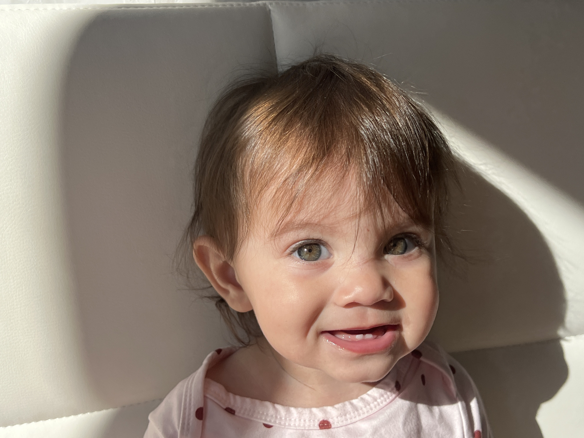 A toddler with light brown hair, hazel eyes, and fair skin is seen from the chest up in a brightly lit indoor scene. The child is wearing a light pink shirt with red polka dots. Their mouth is slightly open, revealing the upper teeth. A ray of sunlight shines diagonally across the background. The setting is unknown.
