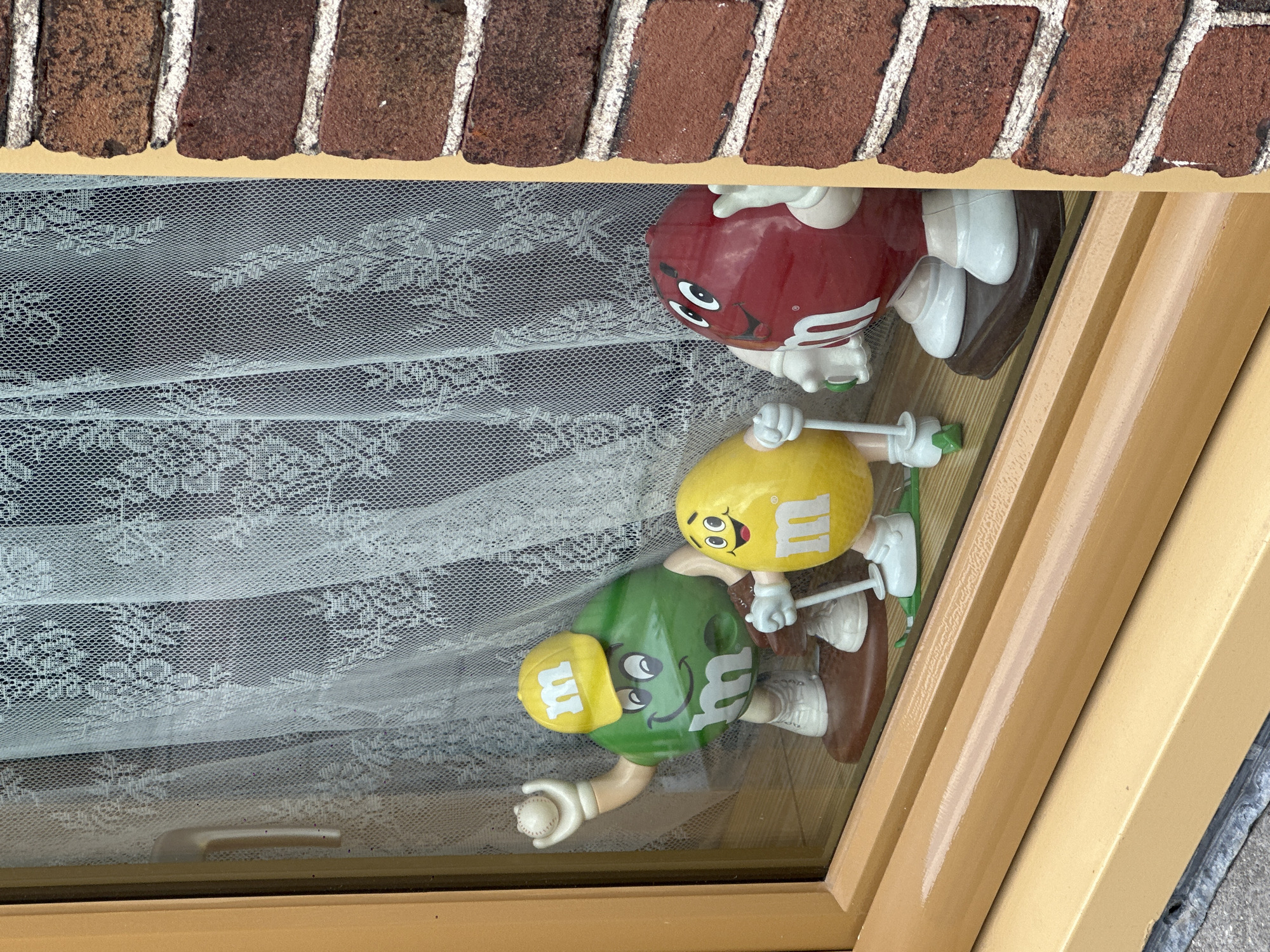 A low-angle shot depicts three M&M candy figures arranged on a surface behind a lace curtain, viewed through a window. The figures are, from top to bottom: a red M&M, a yellow M&M holding a golf club, and a green M&M wearing a baseball cap and holding a baseball. The window frame is painted a golden yellow color, and brickwork is visible above the window. The location is not discernible.
