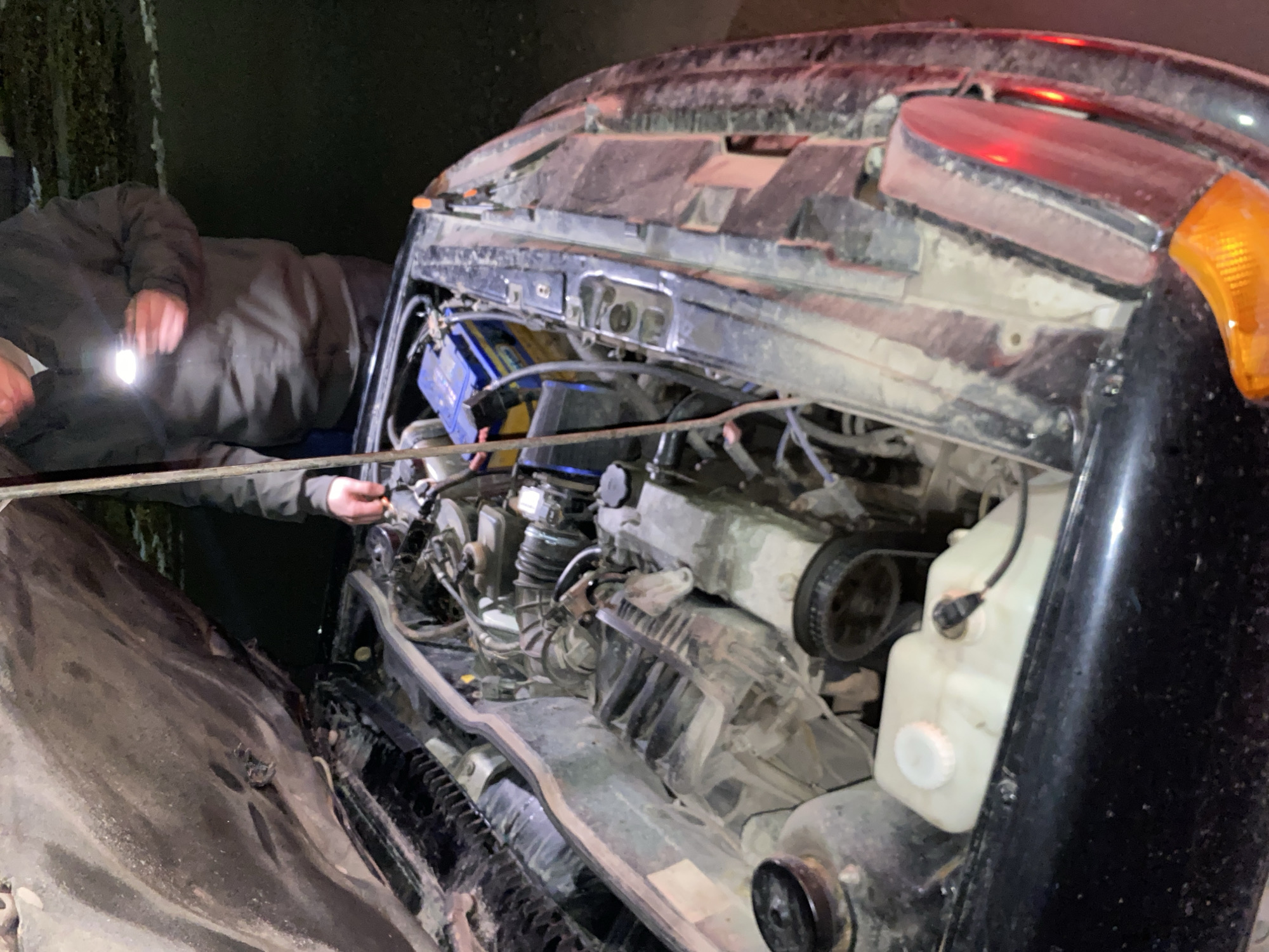 A man is inspecting the engine of a black car. The car appears to be in Chaikovsky, Russia, though this is based solely on context. The engine compartment is open, revealing the engine, battery, and other components. The man is holding a flashlight to illuminate the engine. He is wearing a dark jacket. Another person is partially visible to the left. The car appears to be older and shows signs of wear. The environment is dark.