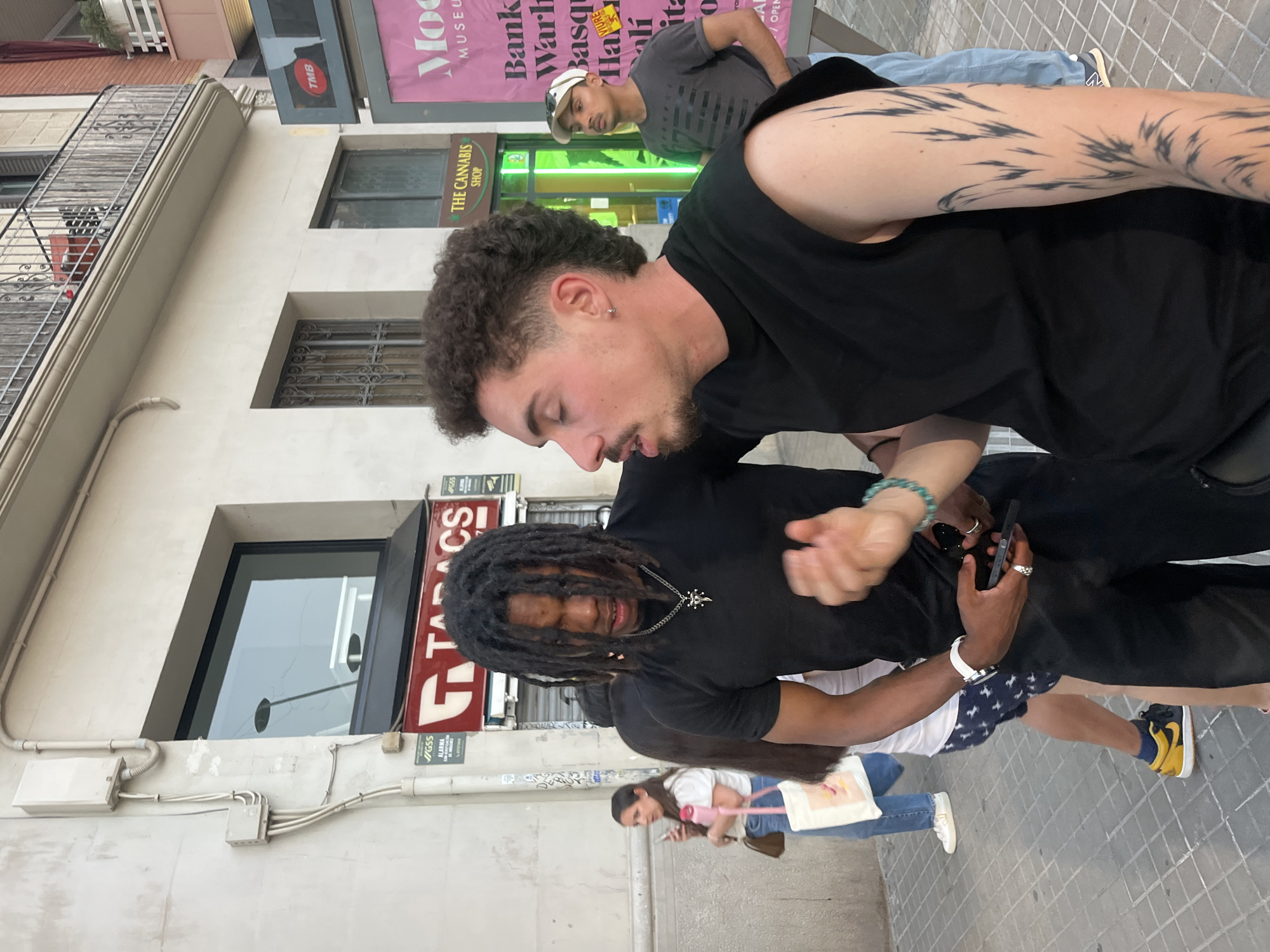 The image shows two young men standing on a city sidewalk in Barcelona, Spain. The man in the foreground on the right has curly dark hair and is wearing a black sleeveless shirt. He has a tattoo on his left arm and is looking to his left. To his left, another man with dreadlocks and wearing a black t-shirt is speaking or looking towards the right. He is wearing a necklace. In the background, other people are visible walking and standing. Behind them, buildings with balconies and windows are present. Signs visible include "TABS" and "THE CANNABIS SHOP." The ground is paved with square tiles.