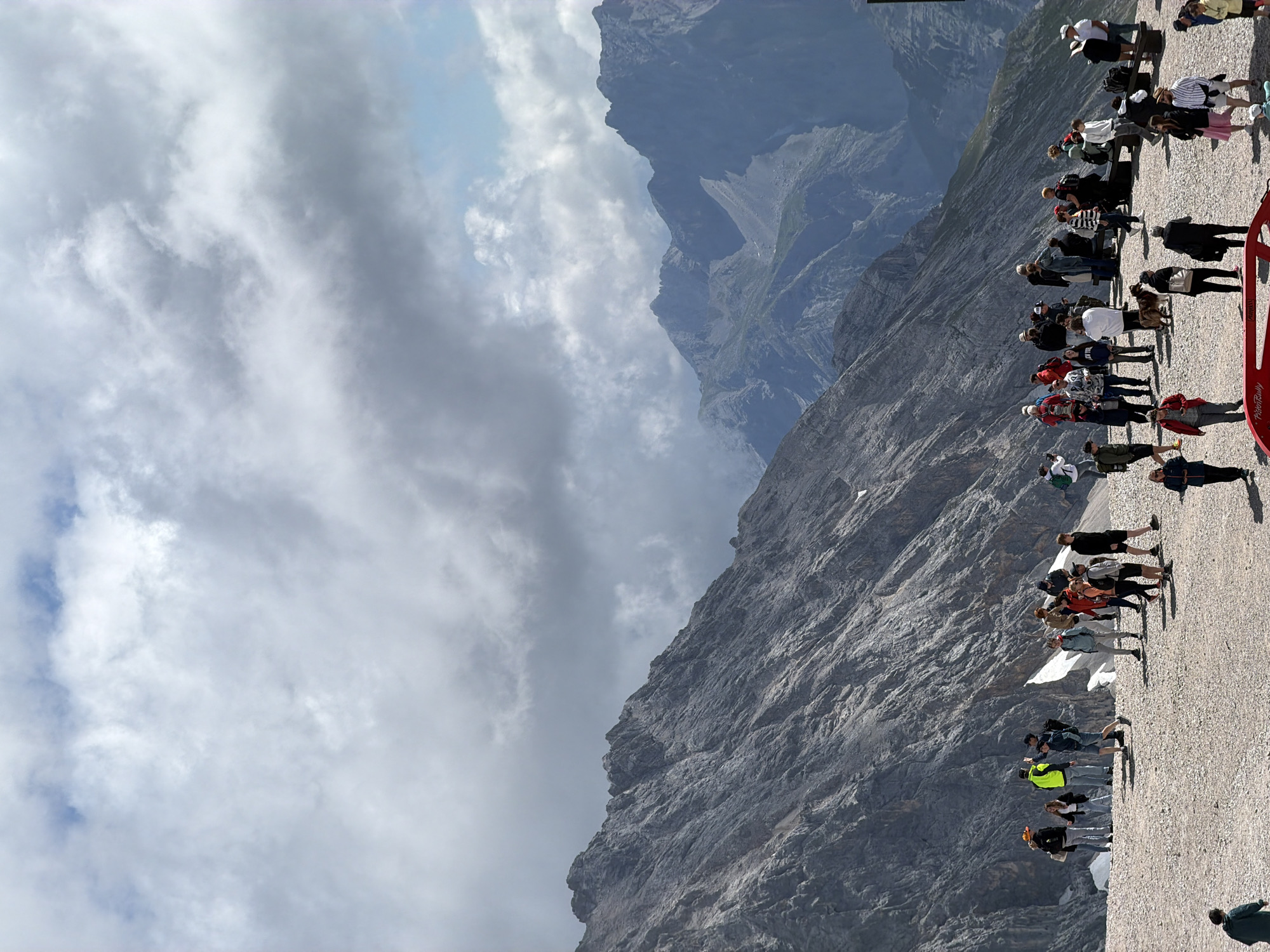 This image depicts a group of tourists gathered on a rocky, mountainous terrain with a vast, rugged mountain range in the background. The sky above is filled with dramatic, heavy clouds, interspersed with patches of blue sky.
The foreground shows a flat, gravelly area where numerous individuals are standing and interacting. They are casually dressed, some with backpacks, suggesting they are on an excursion or visiting a scenic viewpoint. There are several distinct clusters of people, some conversing, others looking out at the landscape, and some seemingly taking photographs. A dog is visible walking among the people.
The mountainous backdrop is characterized by steep, rocky slopes with minimal vegetation. The distant peaks are shrouded in haze, giving a sense of depth and scale to the environment. The overall scene suggests a popular tourist destination in a high-altitude mountainous region, likely accessible to visitors.
Given the context of Garmisch-Partenkirchen, Germany, this scene strongly resembles the summit area of the Zugspitze, Germany's highest mountain. The distinctive rocky terrain, the dramatic mountain vistas, and the presence of numerous tourists are all characteristic of this renowned Alpine destination.