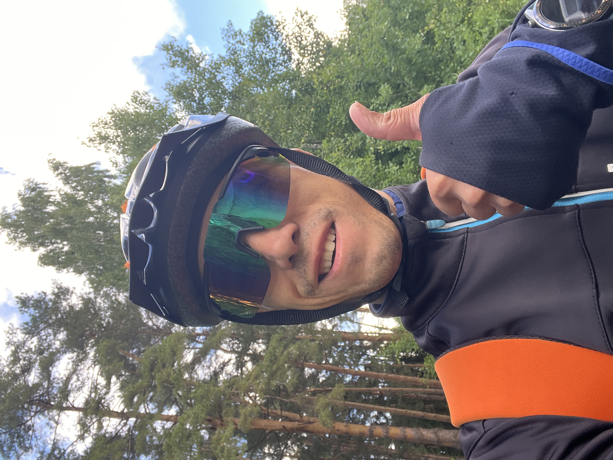 A cheerful selfie captures a man outdoors, likely engaged in an athletic activity. He is shown from the chest up, smiling broadly and giving a thumbs-up gesture with his right hand. He appears to be in his 30s or 40s, with short dark hair and light stubble.
The man is wearing a dark cycling helmet and large, reflective athletic sunglasses with iridescent green and blue hues, which obscure his eyes. He is dressed in a black long-sleeved athletic top or jacket, featuring a prominent bright orange band around the waist and blue piping details around the neckline. A black textured fabric is visible on his right sleeve, and a blue strap, possibly from a watch or another piece of gear, is also seen on his right arm, above a partially visible sports watch.
The background consists of lush green trees, including both coniferous and deciduous types, suggesting a natural, possibly forested, setting. Above the trees, a bright blue sky with some scattered white clouds is visible, indicating daytime with good weather. The sunlight appears to be strong, creating some bright, overexposed areas in the sky. The man's expression conveys happiness, enthusiasm, or approval. The location is Chaikovsky, Russia. No visible text is present in the image.