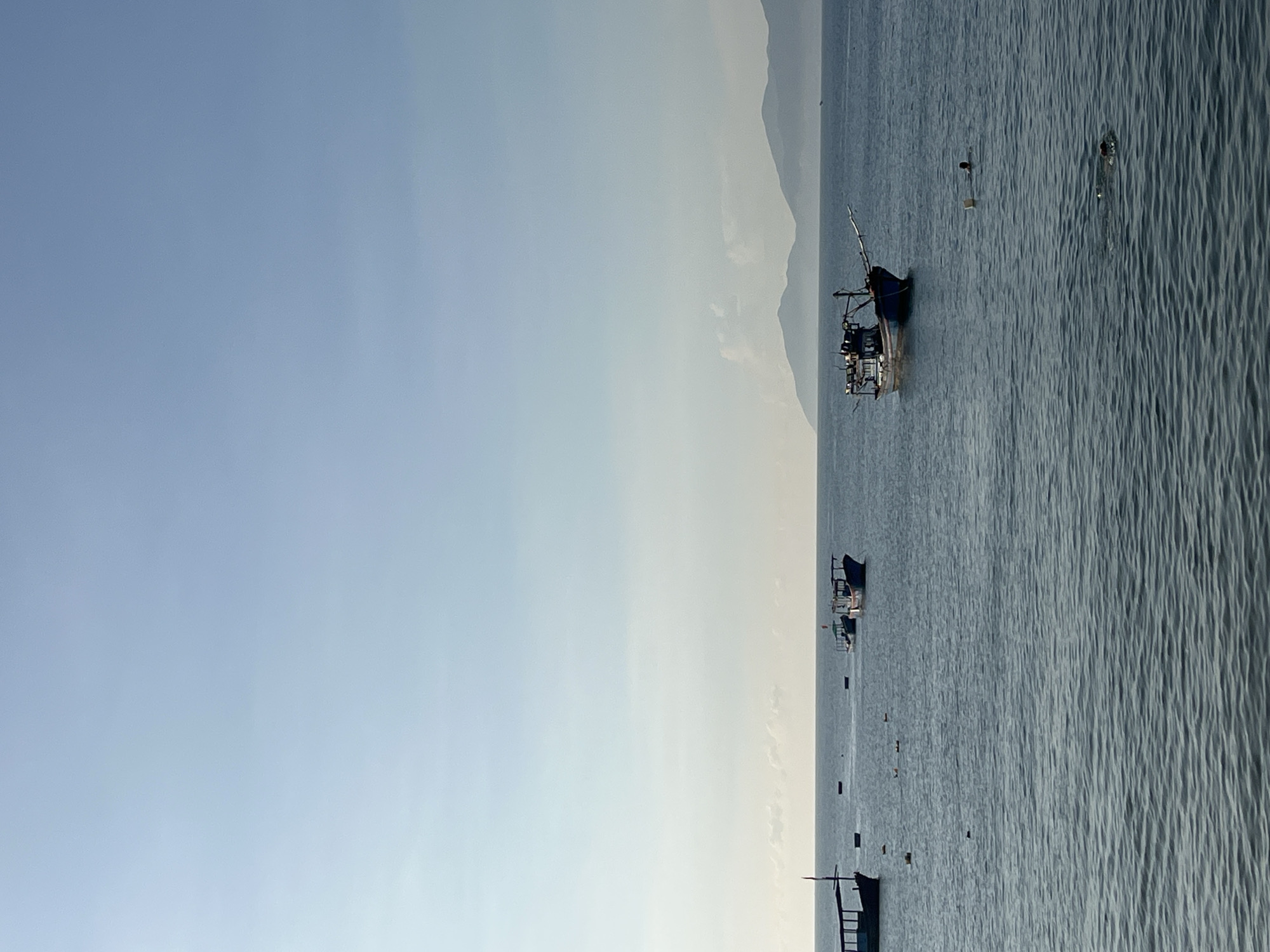 The image shows a body of water with boats and a distant mountain range. The location is Bac Nha Trang Ward, Vietnam. There are several boats of various sizes on the water and some possible humans visible. The sky is pale blue.