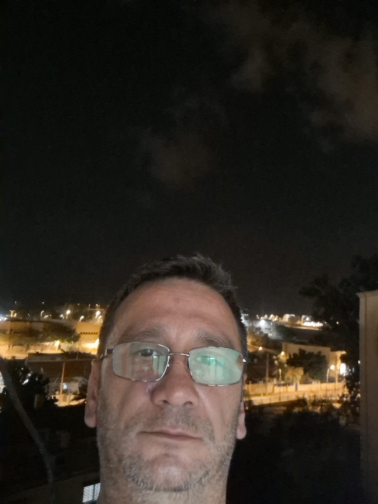 A medium shot selfie taken at night features a middle-aged man with short graying hair and stubble, wearing rectangular eyeglasses. The background shows a blurred cityscape illuminated by streetlights and building lights, likely in Yerocham, Israel, with some trees and structures visible. The sky is dark with some cloud cover. The man's face is close to the camera, with his eyes visible through the reflective lenses of his glasses.