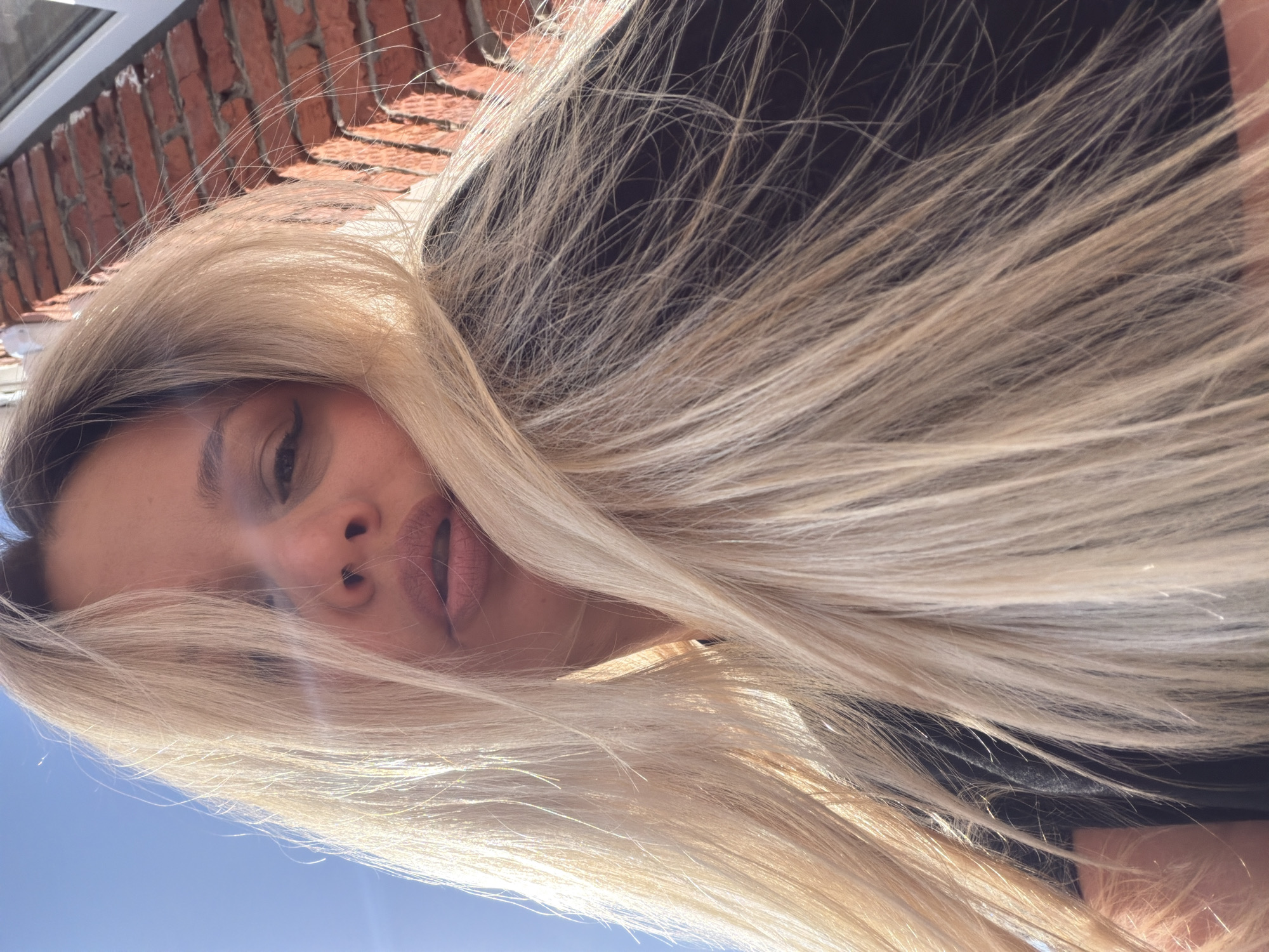 The image is a low-angle selfie of a light-skinned person with long blonde hair. The person's face is visible, including the eyes, nose, and lips. The hair is slightly disheveled and obscures part of the face. Above the person, a brick wall is visible. The sky is seen in the background. There is no indication of the location being Sarapul, Russia.