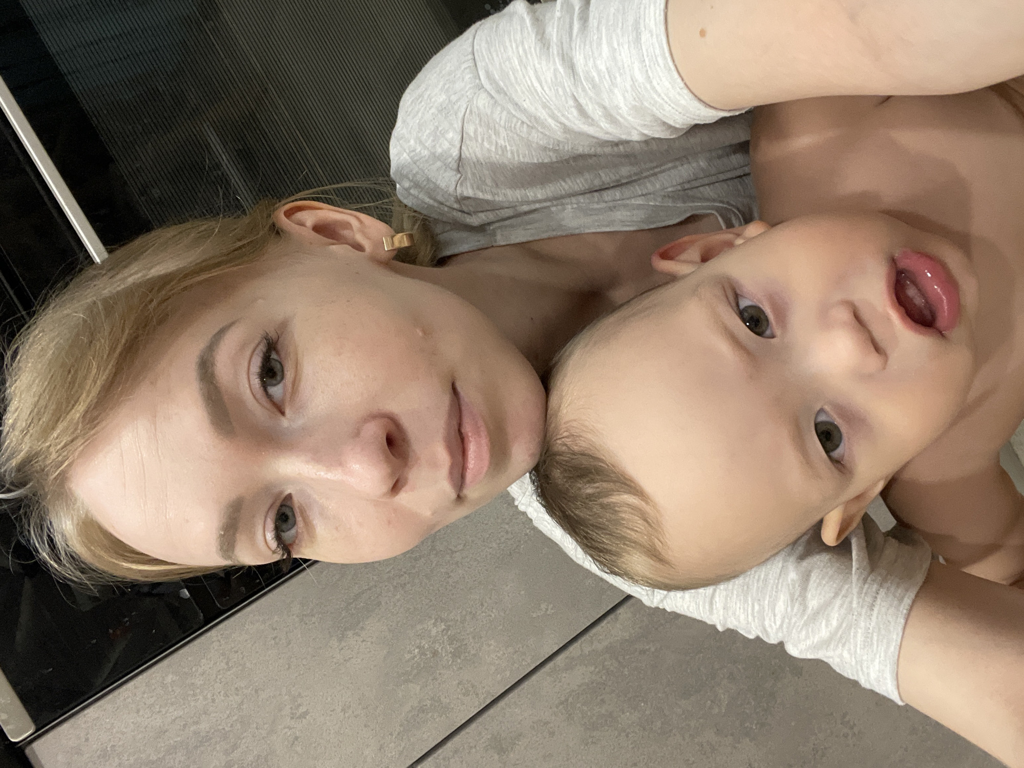 A close-up, rotated selfie shows a light-skinned woman and baby. The woman has light blonde hair, fair skin with minor blemishes, and small gold square earrings. The baby has fair skin, brown hair, and an open mouth, revealing the tongue. Both are wearing light grey clothing. The background includes a dark surface to the left and a light grey surface beneath them. The location and its relation to the city of Perm, Russia, are not determinable from the image.