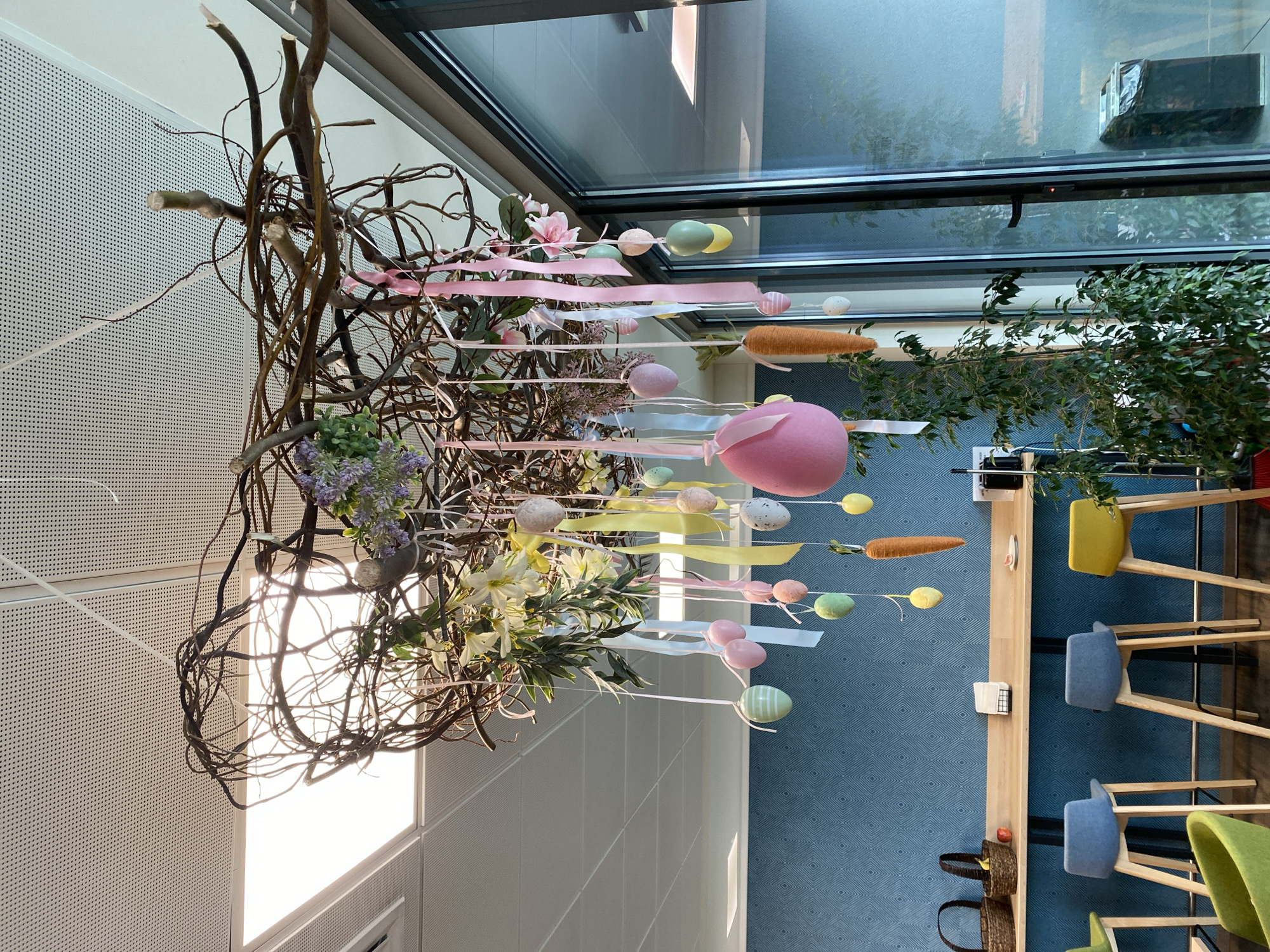 The image shows an interior scene, likely a cafeteria or restaurant, possibly in Sofia, Bulgaria (although this is not definitively confirmed from the image alone). An elaborate Easter decoration hangs from the ceiling, comprising intertwined branches, ribbons in pastel colors (pink, yellow, white), Easter eggs, carrots (likely decorative), and flowers (lilac, white, and pink magnolias).
Below the decoration, there is a long, light-colored wooden counter with several bar stools in varying shades of green, yellow, and blue. The floor has a patterned blue covering, and a plant stands near the counter. Two wicker baskets sit on the floor near the end of the counter.
The scene is well-lit with natural light streaming through large windows, reflecting off the interior surfaces. There are no people visible in the image.