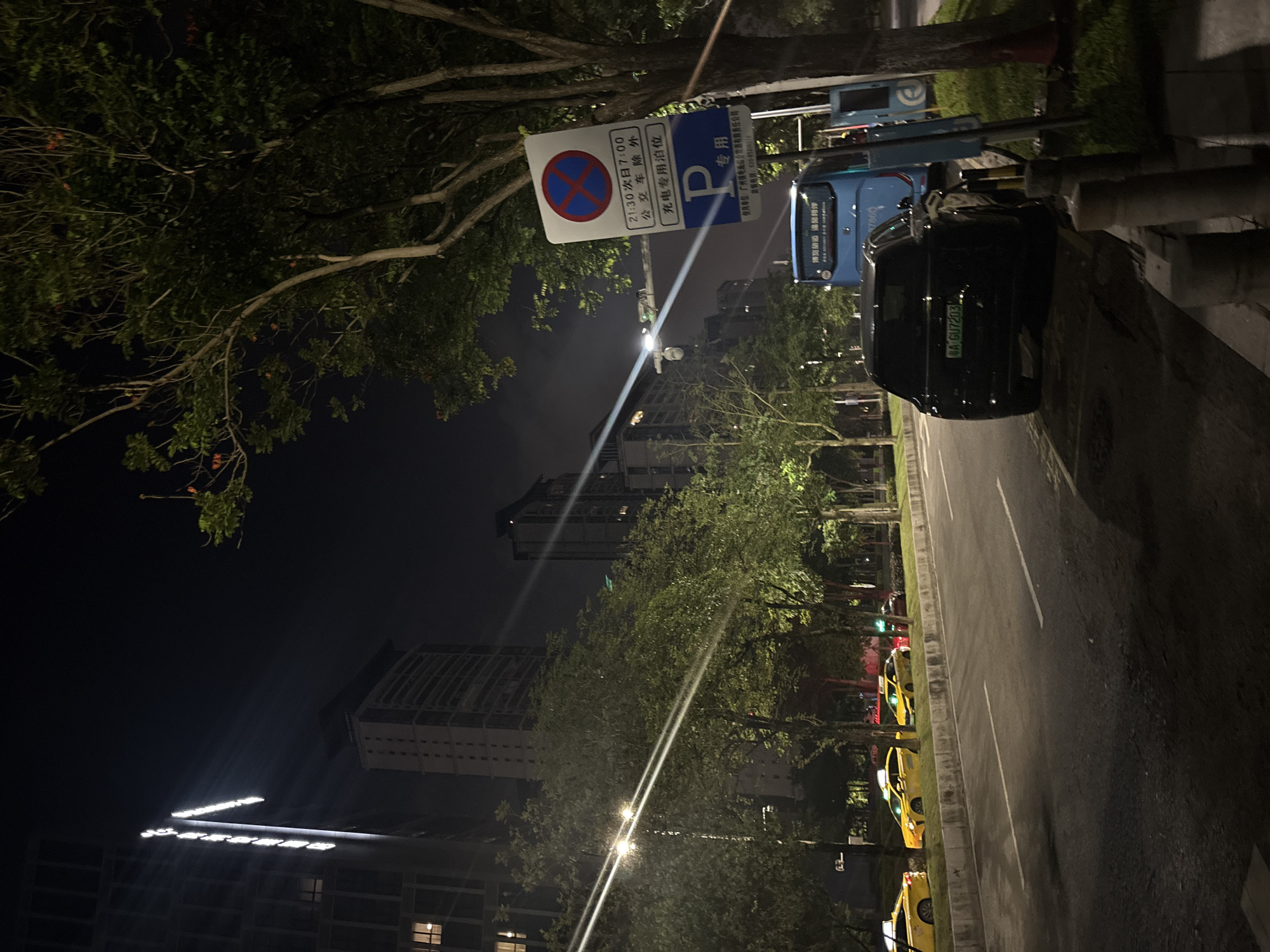 This image depicts a nighttime street scene in the Haizhu District of China. The foreground shows a black SUV parked on the side of a road, with its license plate reading "粤 A GU7203". To the left, several yellow taxis are lined up along the curb. In the background, tall buildings are illuminated against the dark sky, with bright lights emanating from one building's vertical signage. Trees line the street, their leaves illuminated by streetlights. A blue bus is also visible, parked next to a charging station. A traffic sign displaying parking regulations, including times and specific vehicle restrictions, is prominently displayed on the right side of the image. The street is wet, suggesting recent rain. The overall atmosphere is urban and nocturnal.