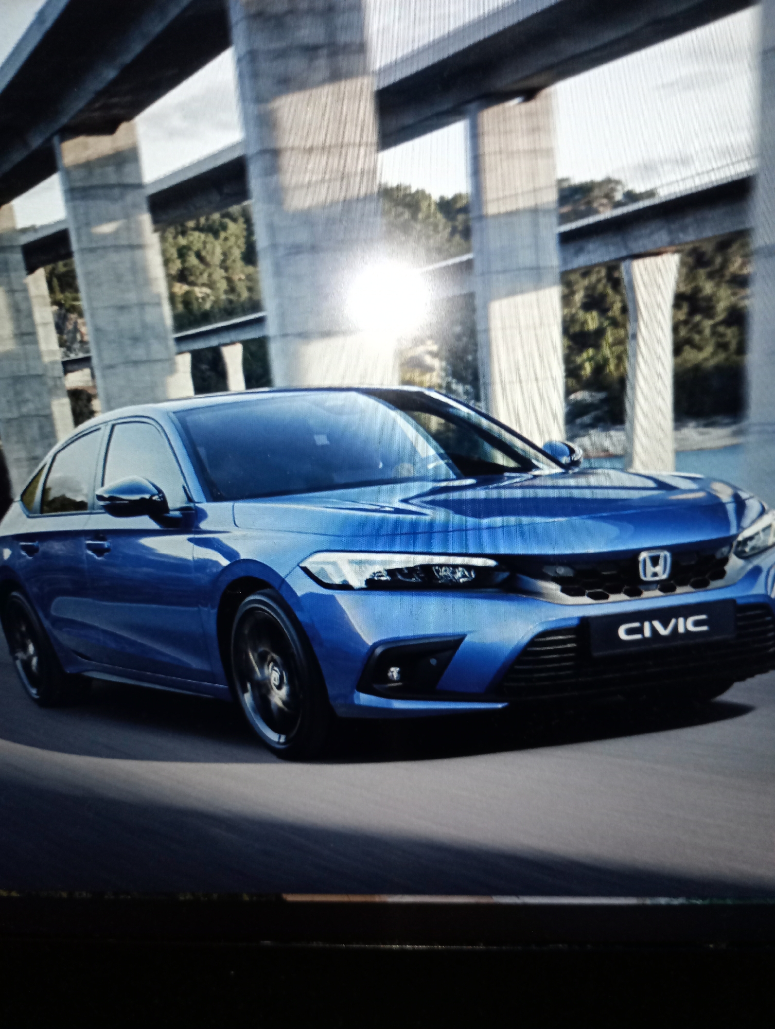 A blue Honda Civic is driving on a road under a large concrete overpass. The car is in motion, and the wheels are blurred. The overpass has large concrete pillars supporting the roadway above. There are trees and hills in the background, suggesting a semi-urban or rural setting. The sun is shining, creating bright reflections on the car and the overpass. The image appears to be a photograph taken from a slightly low angle, emphasizing the car and the imposing structure of the overpass. There are no people visible in the image. The location is not specified to be Harmanli, Bulgaria.