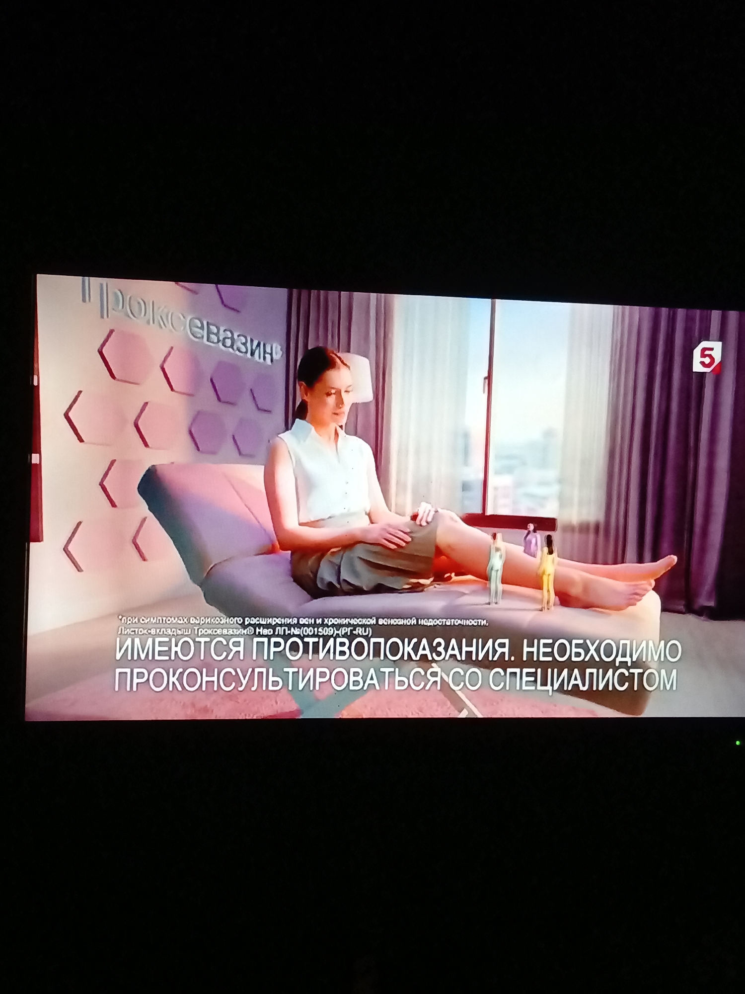The media file is a photograph of a television screen displaying an advertisement. The scene features a woman sitting on a chaise lounge in what appears to be a well-lit, modern room with large windows and purple curtains. The woman is wearing a light-colored sleeveless top and dark trousers, with her legs crossed. In front of her, on the chaise lounge, are three small figurines of people.
Behind the woman, on the wall, are stylized hexagonal shapes in pink and purple, and the word "ТРОКСЕВАЗИН" is visible in large, stylized letters. To the right of the scene, a red logo with the number "5" is visible in a white circle, indicating a television channel.
Text at the bottom of the screen, in Russian, states: "*при симптомах варикозного расширения вен и хронической венозной недостаточности. Листок-вкладыш Троксевазин® №0 ЛП-№(001509)-(PG-RU) ИМЕЮТСЯ ПРОТИВОПОКАЗАНИЯ. НЕОБХОДИМО ПРОКОНСУЛЬТИРОВАТЬСЯ СО СПЕЦИАЛИСТОМ*". This translates to: "*for symptoms of varicose veins and chronic venous insufficiency. Leaflet Troxevasin® No. LP-No. (001509)-(PG-RU) CONTRAINDICATIONS EXIST. CONSULT A SPECIALIST*".
The overall mood is calm and informative, with the advertisement focusing on a medical product. The lighting suggests daytime, and the indoor setting implies a controlled environment. The inclusion of the figurines might symbolize the small scale of the problem or the treatment.