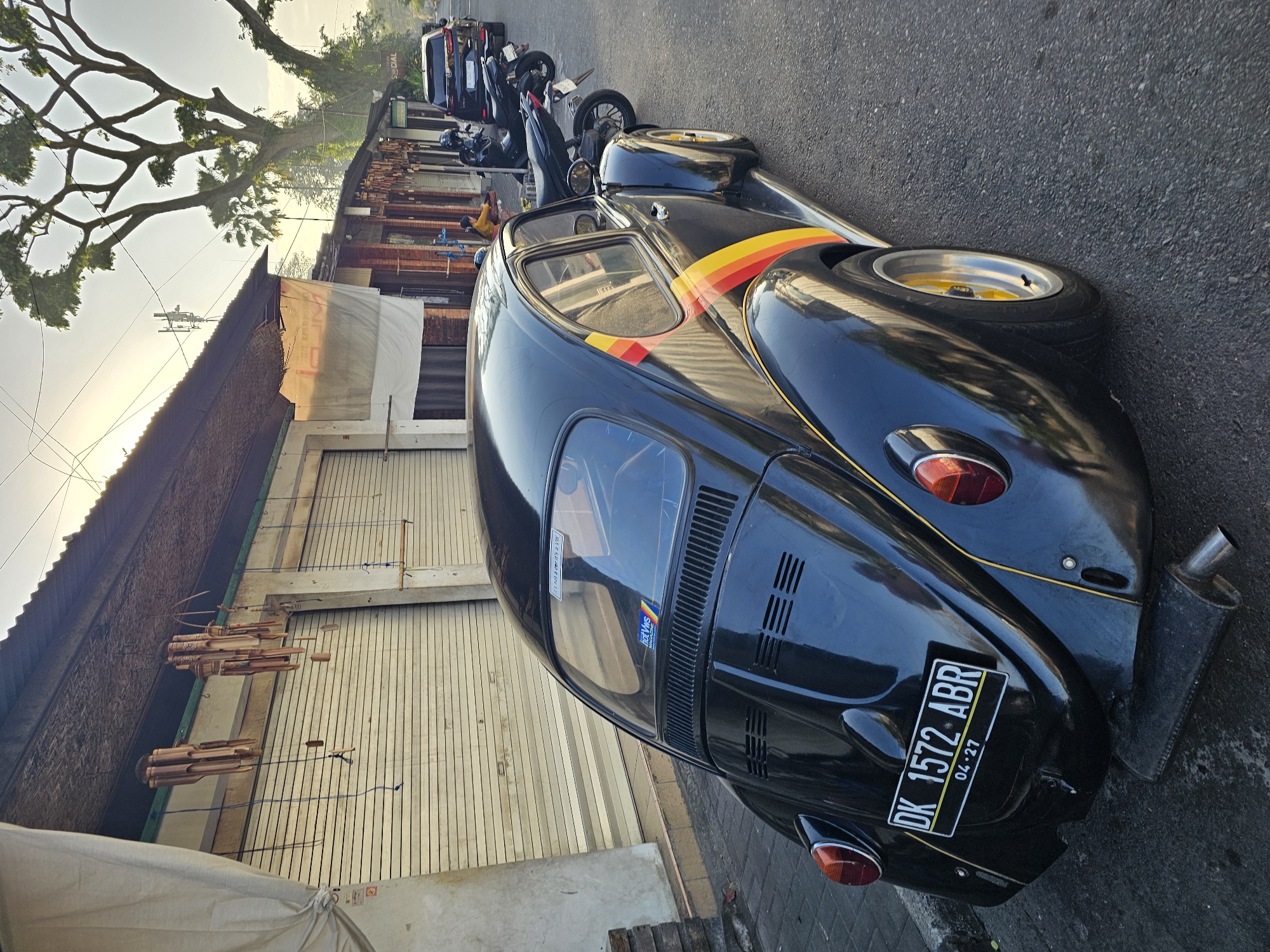 A black Volkswagen Beetle with a yellow and red stripe on the side is parked on a street in Denpasar, Indonesia. The car has a license plate that reads "DK 1572 ABR". In the background, there are several shops with rolled-down shutters and a few motorcycles parked on the side of the road. A woman in a yellow shirt is sitting on one of the motorcycles. The sun is setting, casting a warm glow over the scene. The environment is urban, with a paved road and buildings lining the street. There are trees in the background, suggesting a tropical or subtropical climate. The overall impression is one of a peaceful, everyday scene in a Southeast Asian city.