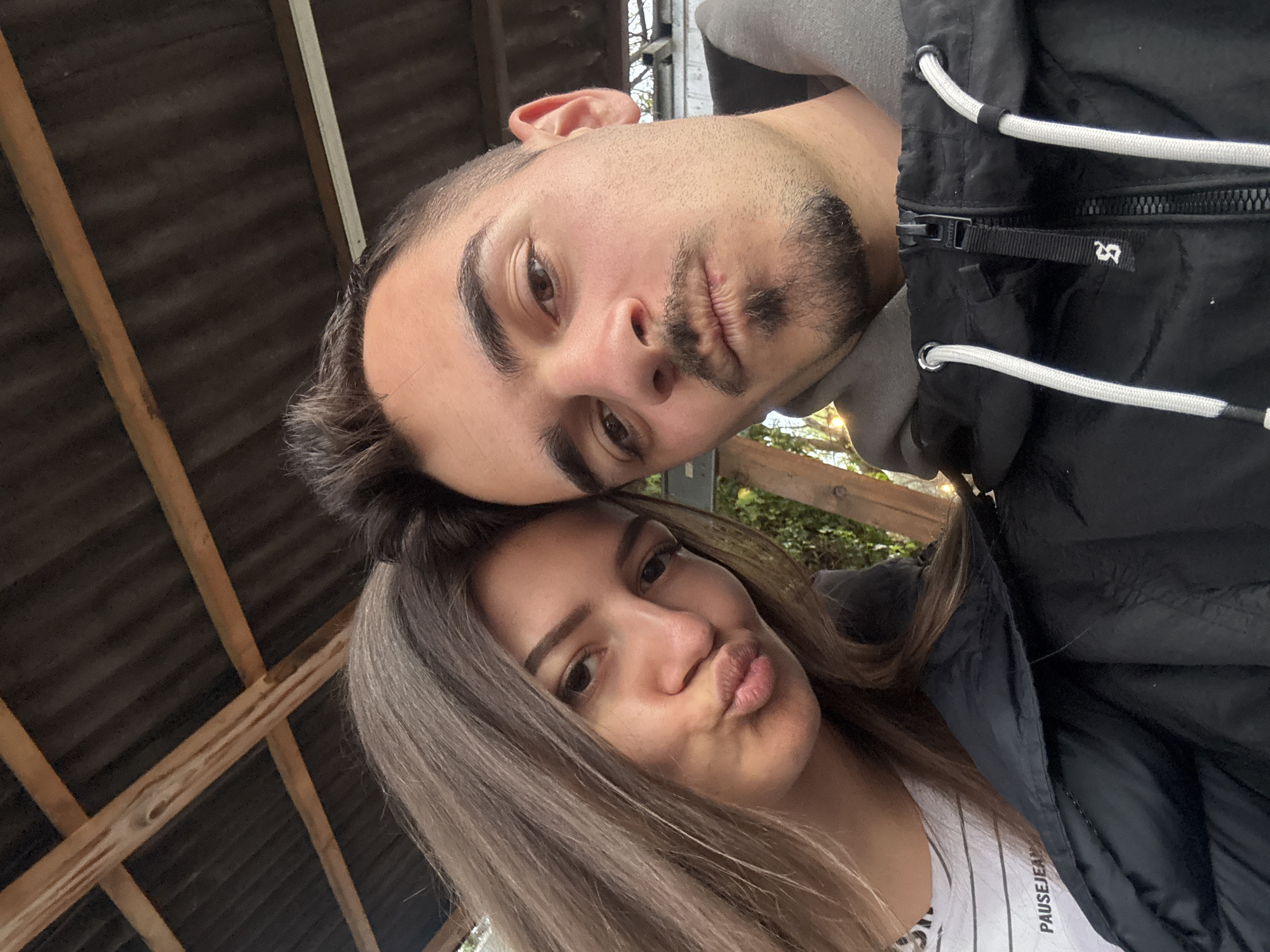 A tilted close-up selfie shows a young man and woman looking directly at the camera, both making a "duck face" expression. The man is positioned above and slightly behind the woman.
The man has short, dark hair and light stubble/goatee. He has dark eyes and is wearing a grey hooded top underneath a black jacket. His jacket features white elastic cords, a prominent zipper, and a small white stylized logo that resembles "2R" or "LR" on the right chest area.
The woman has long, straight light brown hair with subtle highlights. She also has dark eyes. She is wearing a white t-shirt with black horizontal stripes, with the text "PAUSEJEANS" visible on it, layered under a black jacket.
The background suggests an outdoor or semi-outdoor setting, possibly under a shelter or pergola. Above and to the left, a dark corrugated roof and wooden support beams are visible. To the right, some blurred greenery and bright, possibly overcast, sky can be seen, indicating daytime. The scene is consistent with a casual moment in Sofia, Bulgaria.