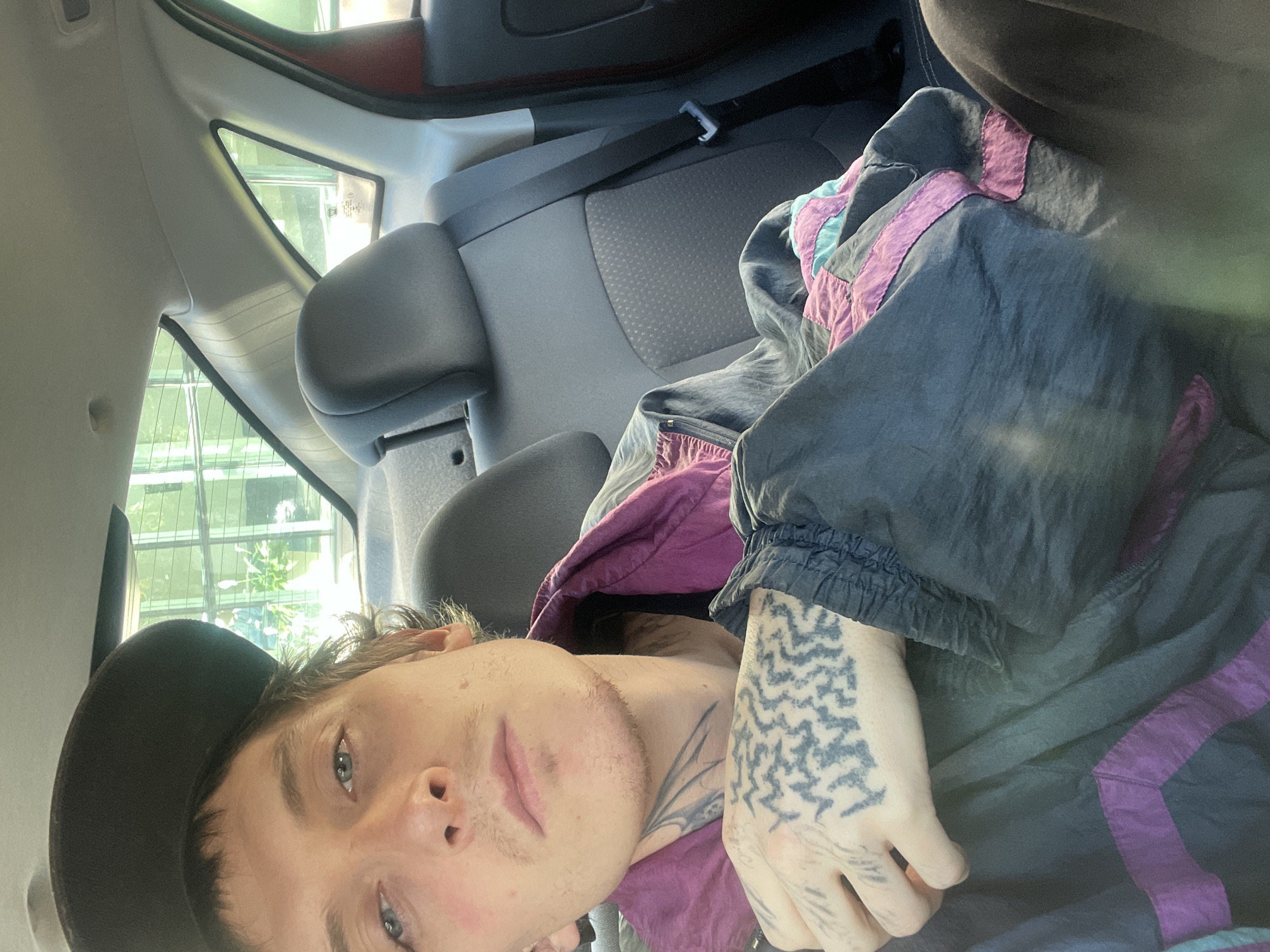 A medium shot depicts a person in the back seat of a car. The individual is lying down, wearing a black baseball cap and a jacket with purple and teal accents. Visible on the person's hand is a tattoo. The car's interior, including the headrest and seatbelt, are visible. A window shows a view of what seems to be a building or complex of buildings with vertical structures. The location is not specifically discernable as Olivos, Argentina, based on the image.