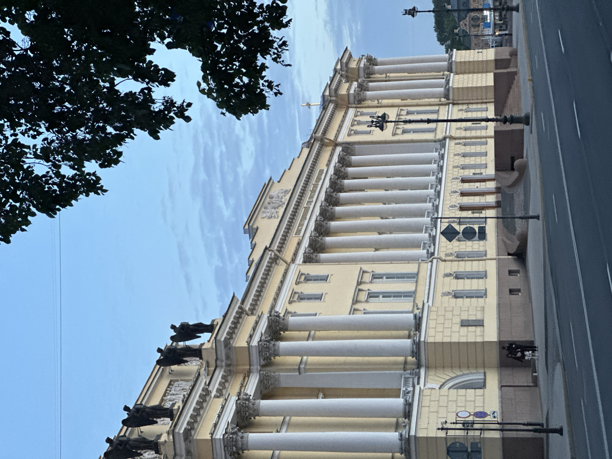 A low-angle, full shot captures a neoclassical building with a pale yellow facade and a colonnade of white Ionic columns. Sculptures of winged figures adorn the top of the building. A large tree with dark green leaves partially obscures the top right corner of the frame. The sky above is a pale blue with wispy clouds. A street with white lane markings runs in the foreground. Two figures, a male and female, stand on the sidewalk in front of the building, conversing. Streetlights and traffic signs are visible along the sidewalk. The environment appears to be urban, likely in the late afternoon or early evening, given the soft light.