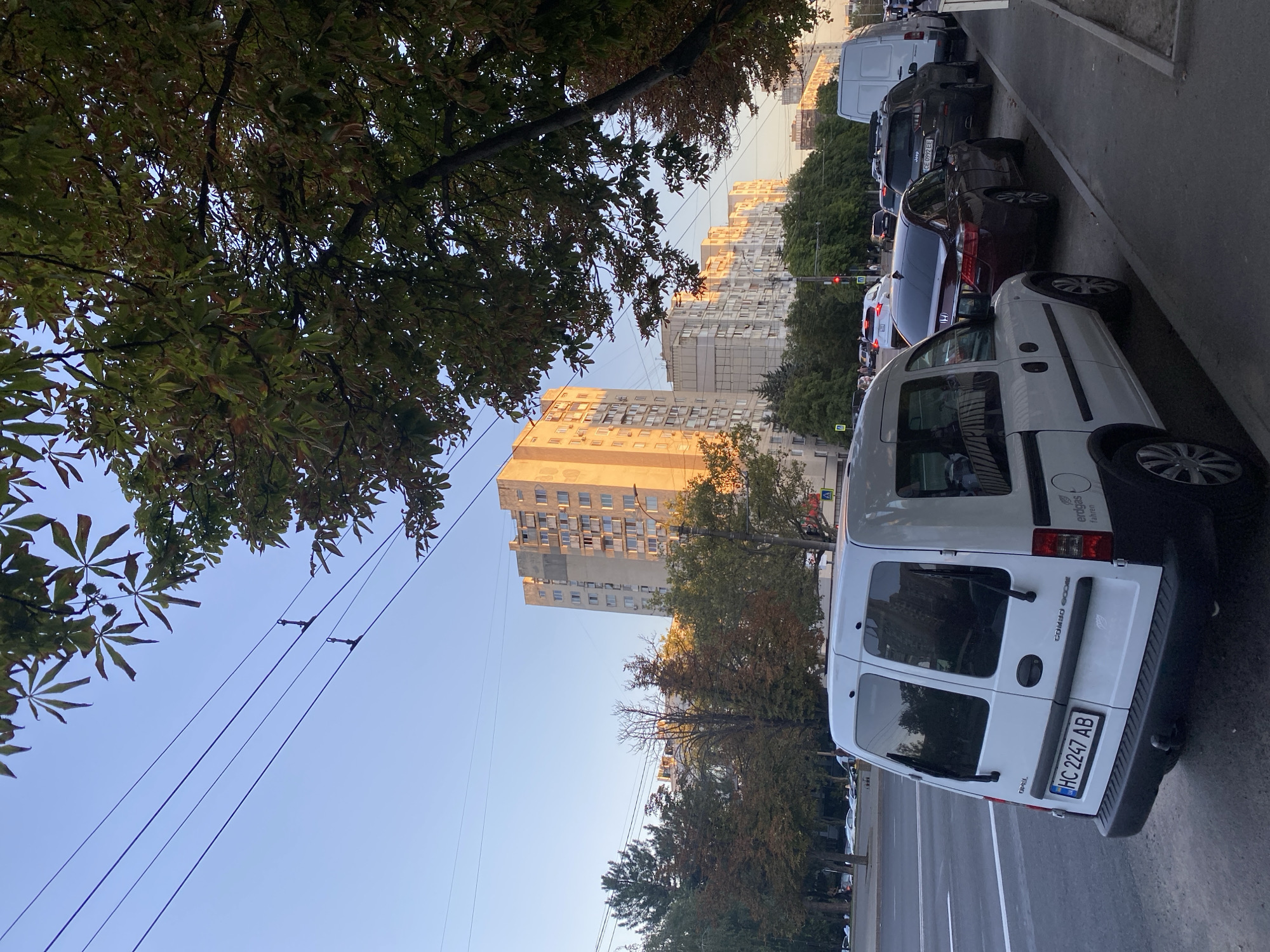 A white Opel Combo van is parked on the side of a street in Chișinău, Moldova, with its rear facing the viewer. The van has a Ukrainian license plate that reads "HC 2247 AB". In front of the van, several other cars are parked along the sidewalk, including a dark red Honda sedan and a black Jeep SUV. Beyond the parked cars, a multi-lane road is visible with traffic. On the opposite side of the road, several multi-story apartment buildings line the street, with trees and power lines in between. The sky is clear and blue, with the sun setting and casting a warm glow on the buildings. The overall scene depicts a typical urban street in Chișinău during the late afternoon or early evening.