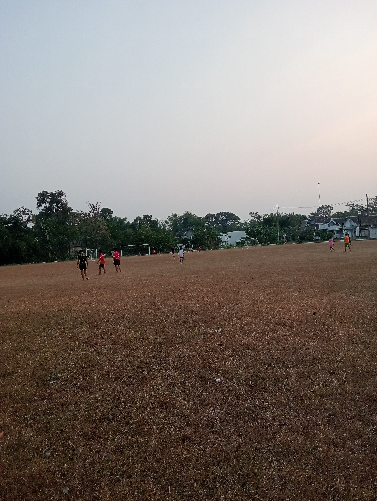 The image depicts an outdoor scene on a dry, brown grassy field, likely a sports ground, at what appears to be late afternoon or early evening. Several individuals, seemingly children and teenagers, are scattered across the field, engaged in or observing a game of soccer. Two soccer goals are visible in the background. The environment is characterized by a hazy, light-colored sky and a line of trees and houses in the distance, suggesting a semi-urban or suburban setting. The overall impression is one of informal recreational activity in a somewhat arid landscape.