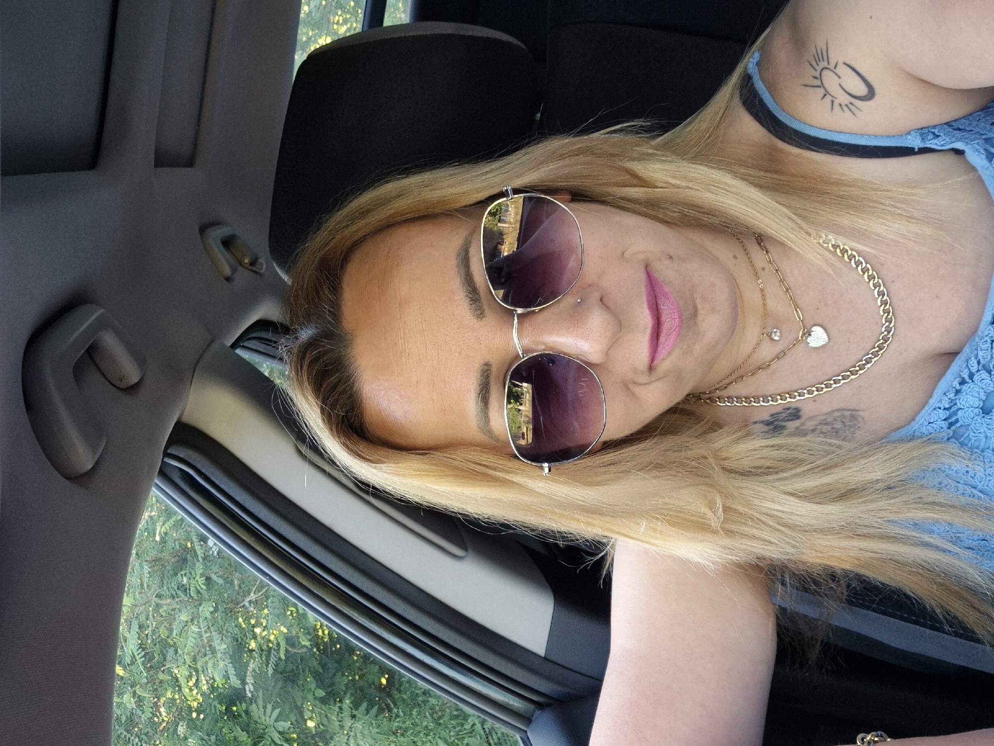 A woman with blonde hair and sunglasses is pictured inside a car. She is wearing a blue top and gold necklaces. A tattoo is visible on her left shoulder. The background shows trees through the car window.