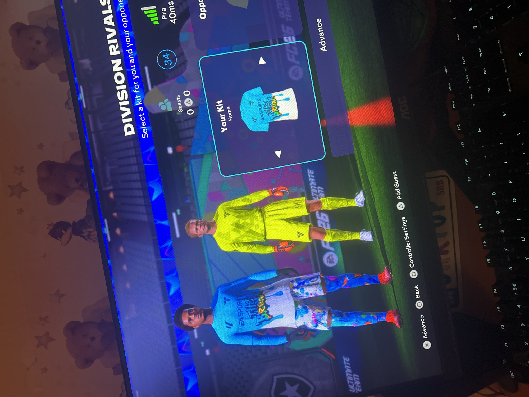 The image displays a screen showing the kit selection screen in the video game EA Sports FC 24. The game is in Division Rivals mode, with the prompt "Select a kit for you and your opponent" visible. The user's home kit is displayed: a light blue jersey with "EA SPORTS THE BRONX" printed on it, along with a colorful graphic. Below the kits are two player models, one wearing the light blue jersey and shorts, and the other wearing a bright yellow goalkeeper kit. On the right side of the screen are menu options, including "Advance", "Back", "Controller Settings", and "Add Guest." The ping is displayed as 40ms.
The screen is being viewed next to a black keyboard, which has a "VERTU SRB" sticker and an "AOC" monitor. There is also a surface visible with a teddy bear and stars pattern. The location of Sofia, Bulgaria is not discernible in this image.