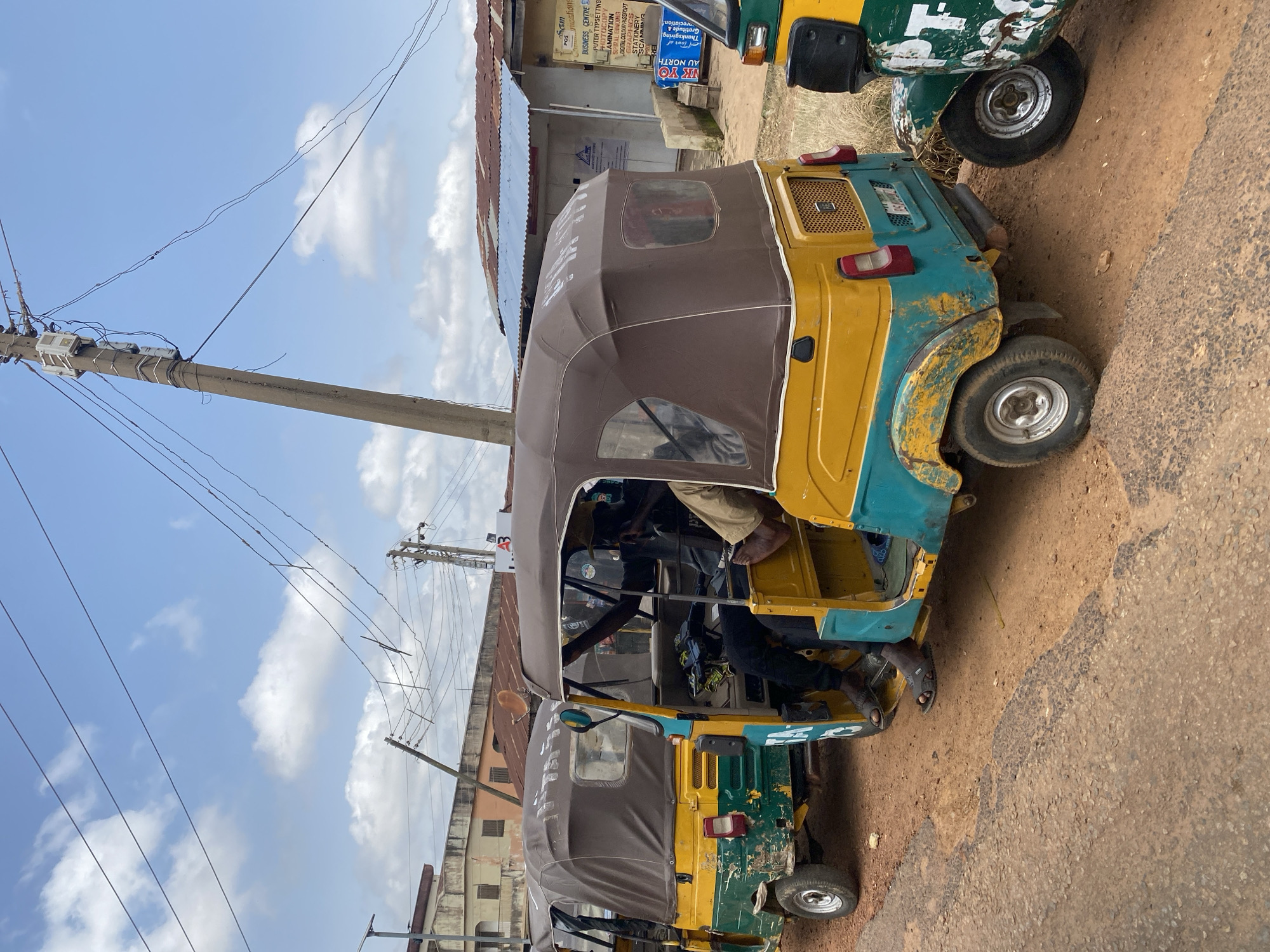 In the city of Gwong, Nigeria, a row of three yellow and teal auto-rickshaws, commonly known as "keke napep," are parked on a dirt road. The auto-rickshaws, which are open-sided vehicles with a covered canopy, are positioned close together. The lead auto-rickshaw is driven by a person wearing a dark shirt and black pants, with their feet clad in dark sandals. The second auto-rickshaw, partially visible, has a driver wearing a tan shirt and dark pants. The third auto-rickshaw is almost entirely out of frame, with only its rear portion and wheel visible. The auto-rickshaws show signs of wear and tear, with chipped paint and some rust. In the background, a single-story building with a corrugated metal roof and weathered walls is visible, along with utility poles and overhead wires. The sky is a mix of blue with scattered white clouds. The ground is a reddish-brown dirt, with some patches of rough pavement at the edge of the road. The overall scene depicts a typical street view in a Nigerian city, with the auto-rickshaws suggesting a mode of local transportation.