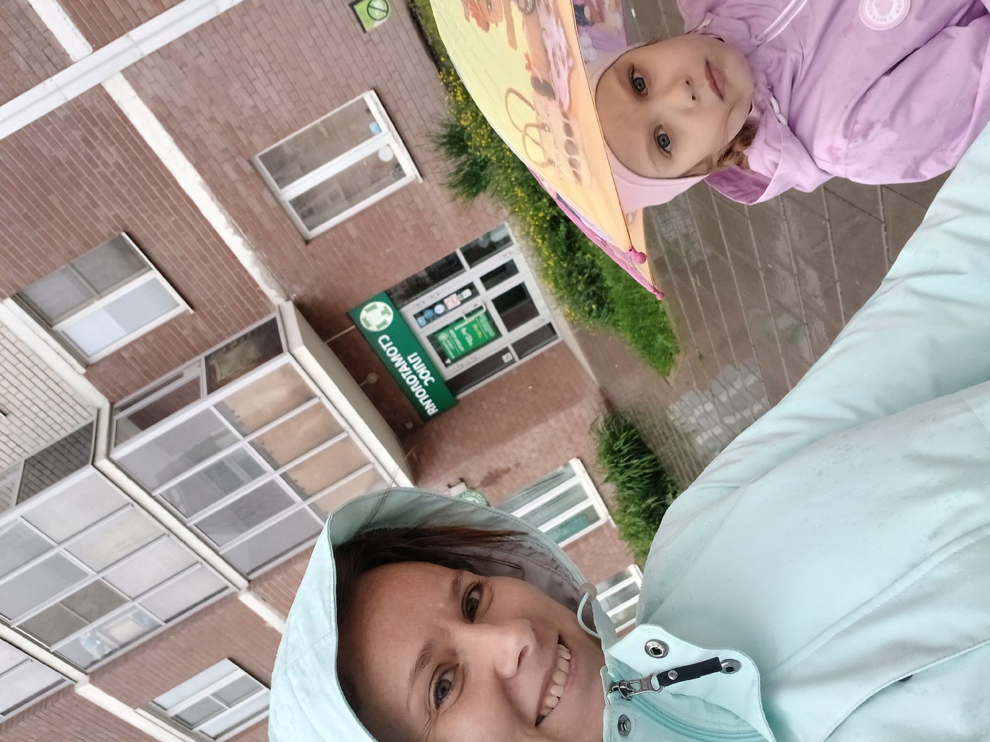 In Yekaterinburg, Russia, on a rainy day, a woman in a light blue hooded jacket takes a selfie in front of a brick building with a glass-enclosed balcony. To her right, a young girl wearing a pink hat and jacket peeks from under a colorful umbrella. The building appears to house a dental clinic, indicated by a green sign with a white tooth logo and text in Cyrillic. The ground is wet, suggesting recent rainfall, and the overall atmosphere is casual and familial, despite the inclement weather.