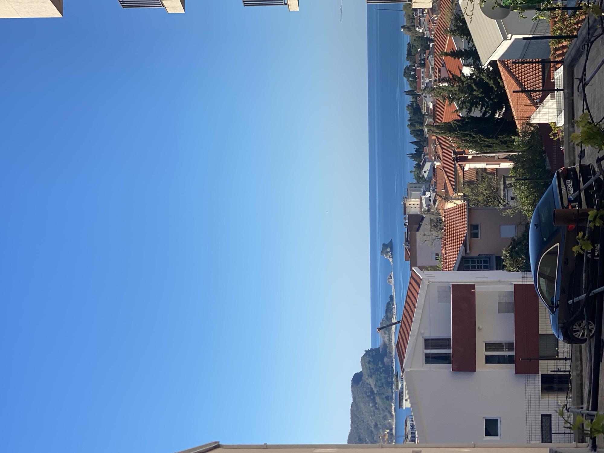 The image shows a view of Budva, Montenegro, from an elevated perspective. The Adriatic Sea stretches towards the horizon, with several small islands visible in the distance. The foreground is dominated by buildings with red tile roofs, typical of the region. A white house with red window shutters is prominent. A black car is parked on the street to the right. The sky is clear and blue. The scene is bathed in bright sunlight.