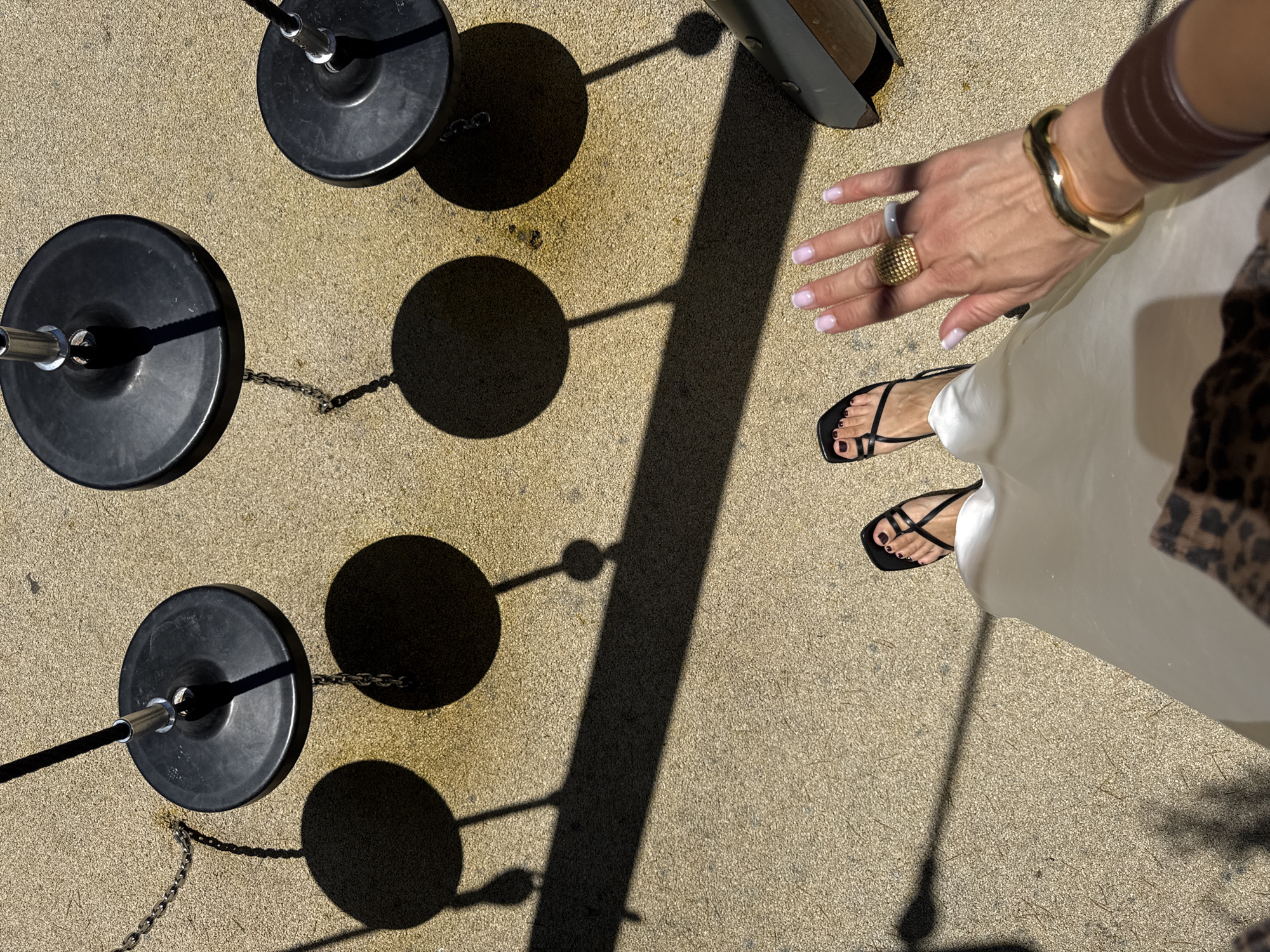An overhead perspective shot captures a woman's feet and hand resting on a sandy-colored outdoor surface, possibly a park or playground, in Sochi, Russia. Four circular black weights, likely part of playground equipment or outdoor exercise apparatus, are visible, casting distinct shadows. The woman's left foot is adorned with black strappy sandals, and her right hand displays manicured nails with a light pink polish, accessorized with a gold textured ring, a grey stone ring, a thick gold cuff bracelet, and a brown cuff. A portion of her cream-colored skirt and a leopard-print garment are also in frame, suggesting a casual, outdoor setting.