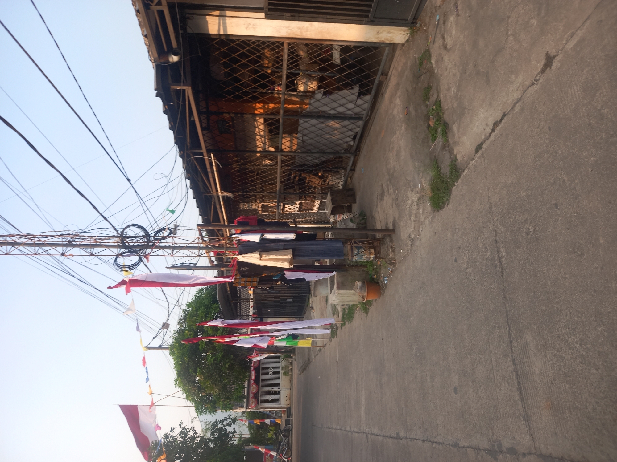The image depicts a street scene in Cankuang, Indonesia, likely during a patriotic celebration, given the prominent display of Indonesian flags and festive banners. The foreground shows a paved road with a slight incline. To the right, a building with a corrugated metal roof and a barred facade is visible, with various items, possibly clothing, hanging on a rack outside. Several Indonesian flags, red and white, are planted along the roadside and also suspended on wires. A telecommunications tower with a tangle of wires stands tall behind the building. In the background, a white fence with decorative elements and gates is seen, along with trees and other buildings. The overall environment appears to be a residential or mixed-use area. The time of day is likely late afternoon, judging by the long shadows.