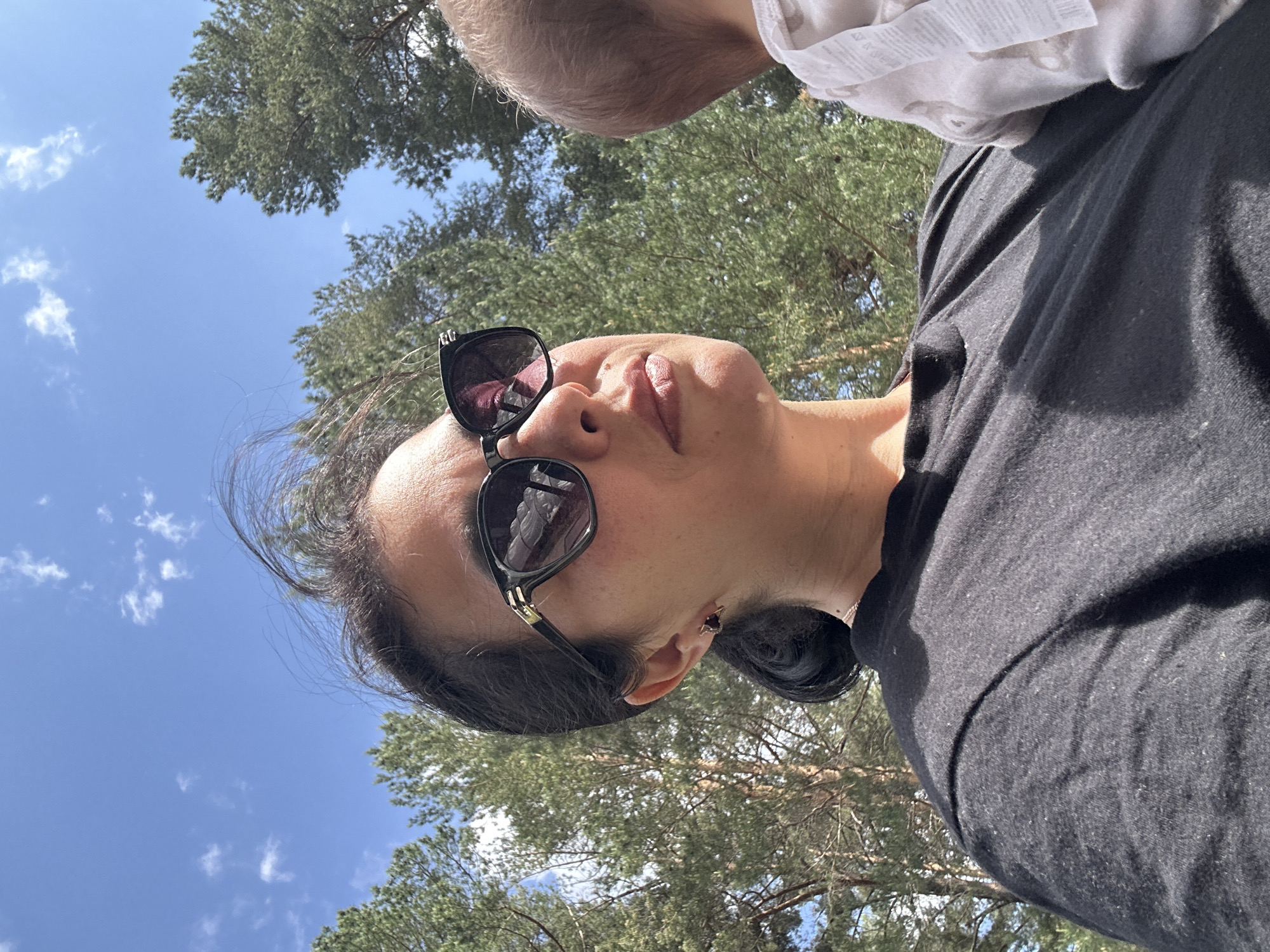 A low-angle, eye-level shot looking up at a person wearing sunglasses and a black shirt. Another person, presumably a child, is partially visible at the top of the frame wearing a white shirt. The background consists of trees and a blue sky with scattered clouds, suggesting the photo was taken outdoors in a wooded area. The city is unknown.