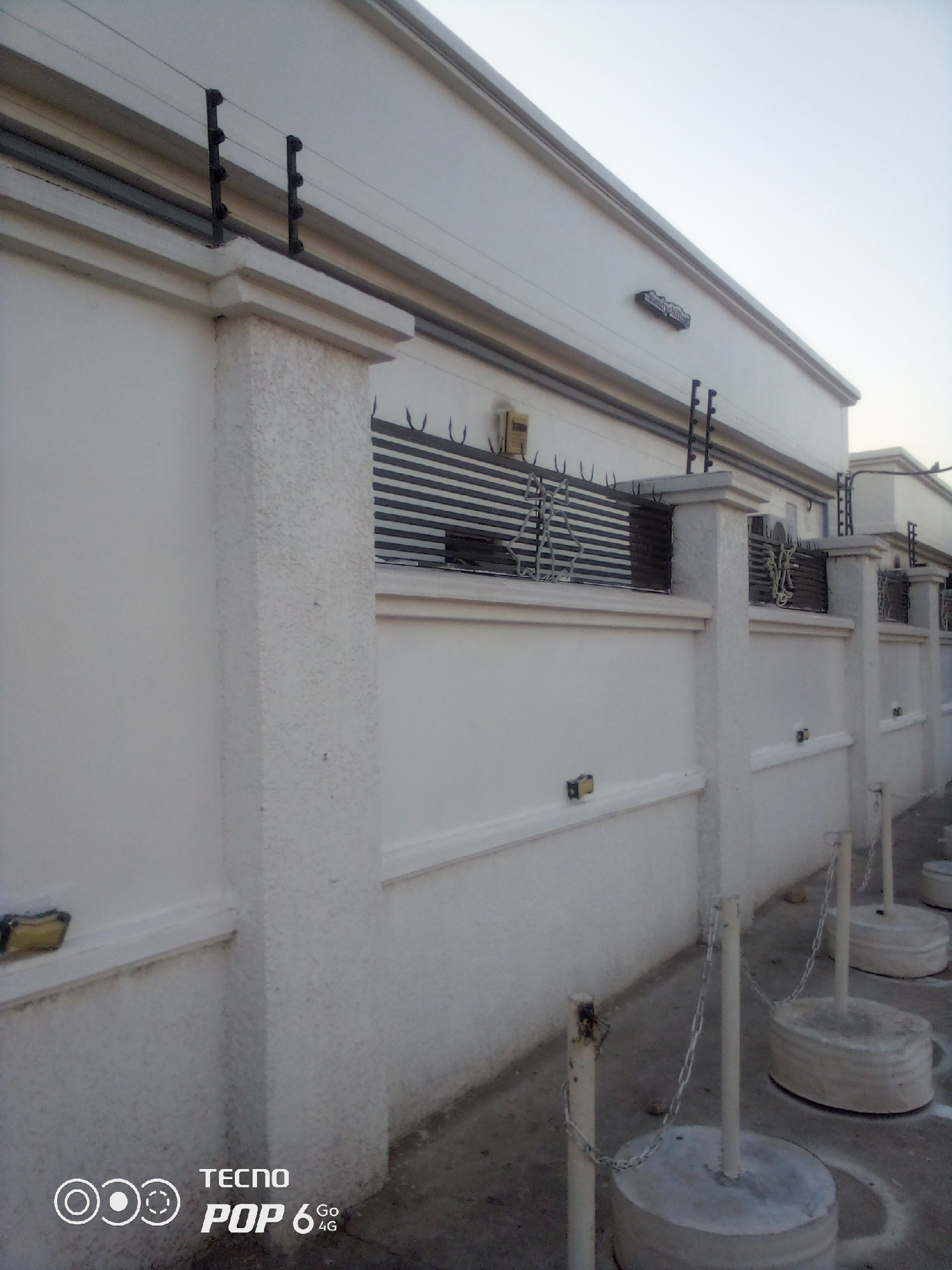 The image depicts the exterior of a building's perimeter wall, likely in a residential or commercial area of Howayi, Nigeria. The wall is constructed of white concrete and features decorative elements and security measures. Atop the wall, an electric fence is visible, with black posts and multiple strands of wire. The wall itself is topped with a horizontal band, above which is a decorative metal grating with pointed spikes.
The wall is punctuated by large, square concrete pillars that support the structure. Between these pillars, the white wall continues, with a horizontal decorative ledge running along its length. Mounted on the wall, at intervals, are small, yellow electrical boxes.
In the foreground, a concrete pavement or walkway runs alongside the wall. Several white metal poles are embedded in the ground, each topped with a circular concrete base. Metal chains connect these poles, suggesting a demarcation or crowd control barrier.
The sky above is a pale, overcast blue, indicating either early morning or late afternoon. The overall impression is one of security and understated architectural design. There are no people visible in the image. The presence of the electric fence and the concrete barriers suggests a focus on security. The TECNO POP 6 4G watermark in the lower-left corner indicates the device used to capture the image.