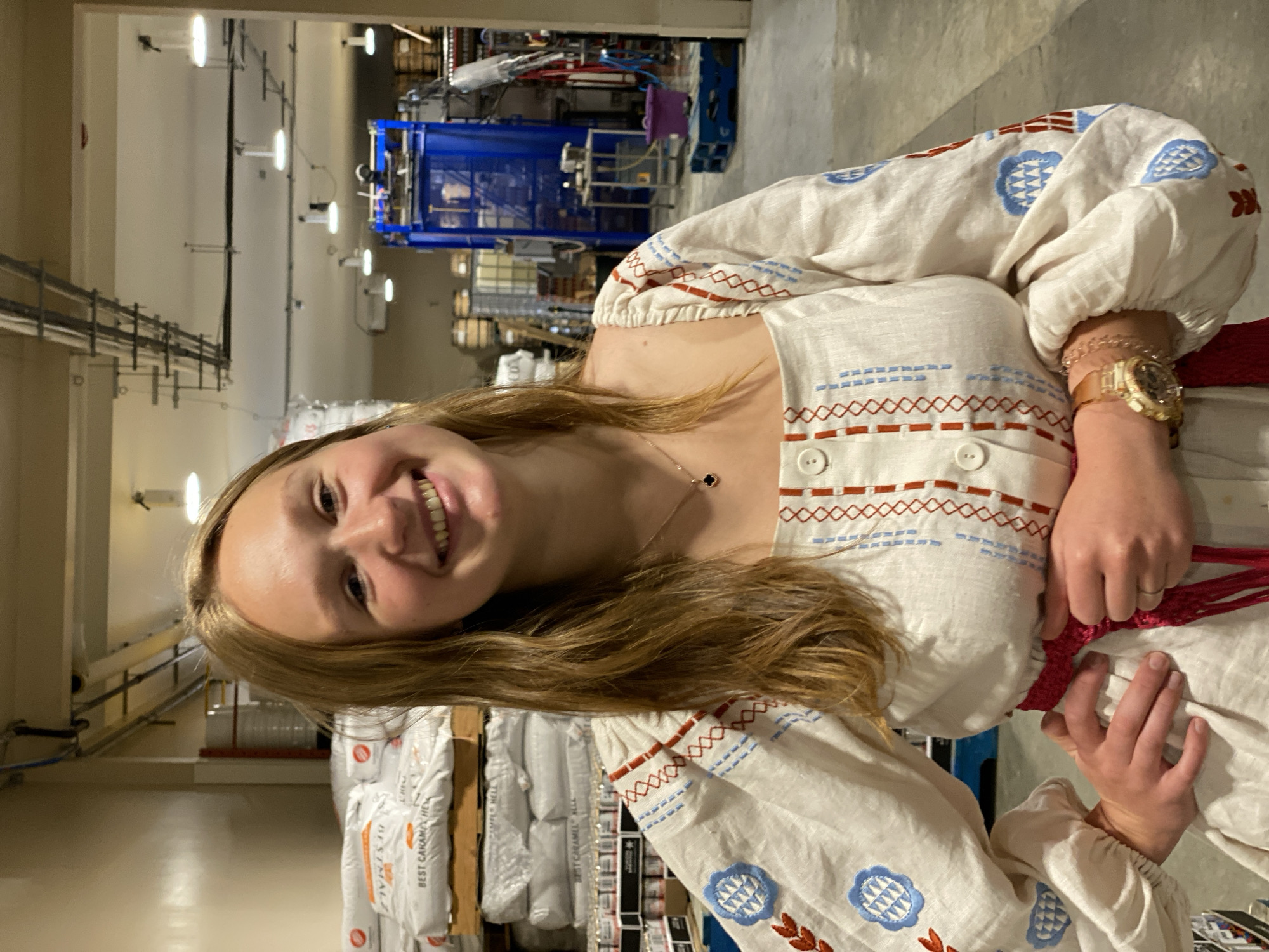 A young woman with blonde hair and fair skin is standing in what appears to be a warehouse or industrial facility. She is smiling directly at the camera, with her left hand on her hip and her right hand resting on her waist. She is wearing a white linen dress with embroidered details in red and blue, a red sash around her waist, and a watch on her left wrist. In the background, there are stacks of large white bags, industrial machinery, and overhead lighting. The environment suggests a place of production or storage, possibly related to food or beverage, given the labeling on the bags that are partially visible. The overall impression is a candid portrait within an industrial setting. There is no specific information to suggest this location is in Whitehorse, Canada.