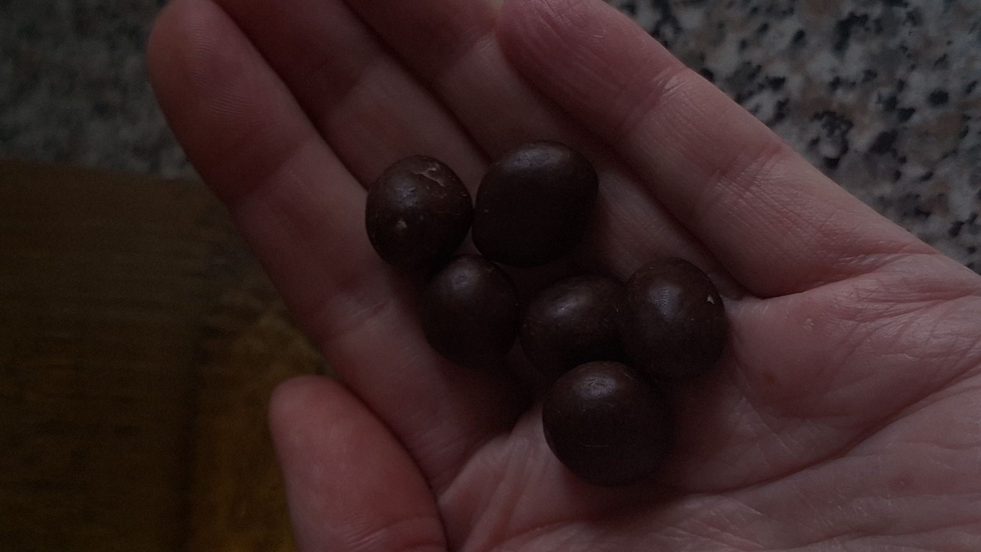 A close-up, overhead shot shows a person's left hand cupping six round, dark brown chocolate-covered candies. The candies are clustered together in the palm of the hand, with some overlapping. The hand is lightly colored with visible fingerprints and lines, resting on a textured, speckled surface, possibly a countertop. In the upper right background, out of focus, is a glass with a golden liquid, suggesting a beverage. The image is lit from above, casting subtle shadows on the candies and the hand. The scene is likely indoors, given the countertop texture. There is no information to determine the specific location in Myadzyel, Belarus.