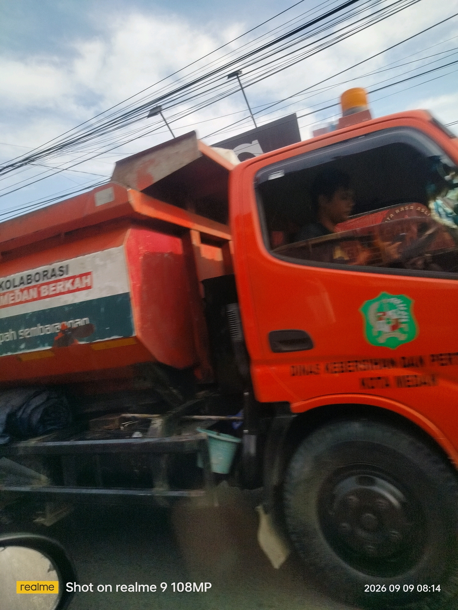 An orange dump truck, identified with text that reads "DINAS KEBERSIHAN DAN PERTA KOTA MEDAN" (Department of Cleanliness and Environment, City of Medan), is driving on a street under a cloudy sky with visible power lines. The truck has a prominent orange cab with a driver visible inside. On the side of the dump bed, there is text that includes "KOLABORASI MEDAN BERKAH" (Collaboration Medan Blessed) and "sampah sembarangan" (littering). A green and white emblem is also visible on the side of the cab. The scene appears to be in motion, likely captured from another moving vehicle. A water mark at the bottom indicates the image was taken with a "realme 9 108MP" camera on "2026 09 09 08:14".