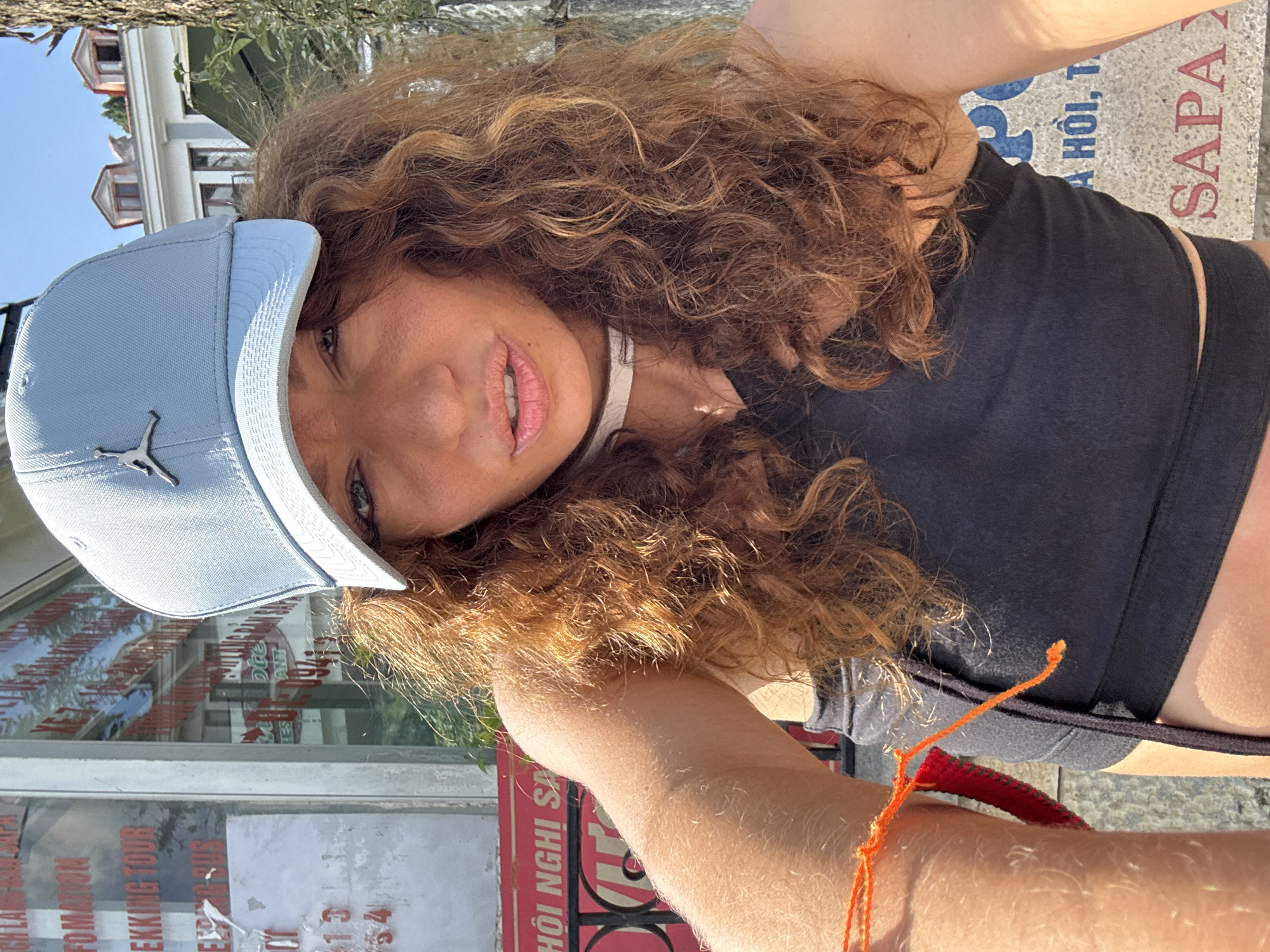 A medium close-up shot captures a woman wearing a light blue Jordan cap with the logo visible, black short crop top and shorts, and a white choker necklace. Her hair is a curly reddish-brown color. She is wearing an orange string tied to her wrist. The woman's face is slightly turned to the side. The background reveals that she is in Sa Pa Ward, Vietnam. Behind her, a glass window shows reflective images of buildings. The text on the glass shows information, including "TREKKING TOUR", "A HỒI", and "SAPA".