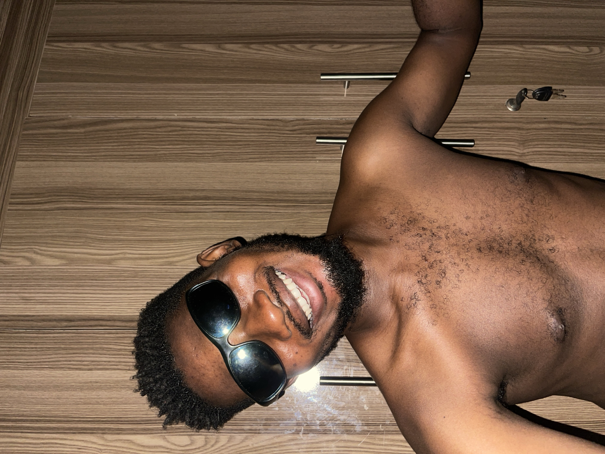 A shirtless, dark-skinned man with a beard and sunglasses lies on his side on a wood-paneled surface. He is smiling. A set of keys rests on the wood-paneled surface near his shoulder. The location is not identifiable as Unguwar Gabas, Nigeria, from the provided image.