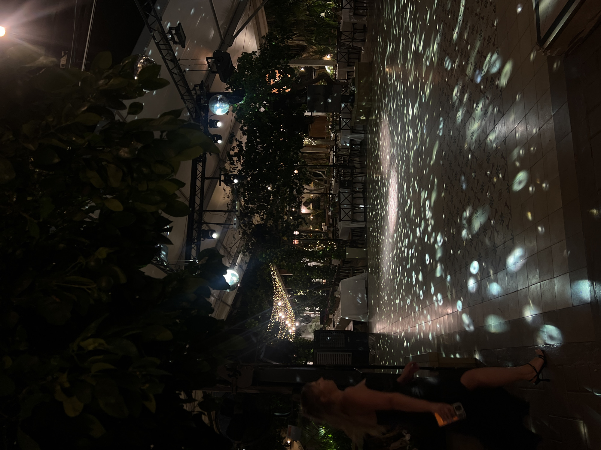The image depicts a dimly lit outdoor event space in Beit Hanan, Israel. A woman in a black dress and high heels stands on the left, holding a drink. The ground is tiled and illuminated by scattered light patterns, likely from disco balls or similar lighting effects. In the background, tables are set for a formal event, with white tablecloths and chairs. String lights are visible in the mid-ground, and large trees with foliage cast shadows. The overall atmosphere suggests a nighttime gathering or party.